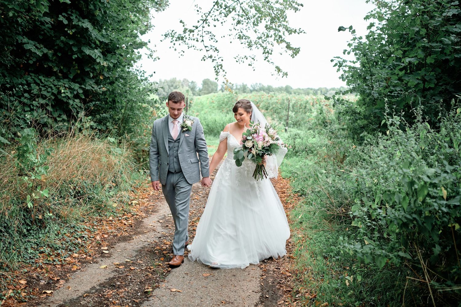 Wedding couple walks together on rustic path surrounded by green summer foliage.