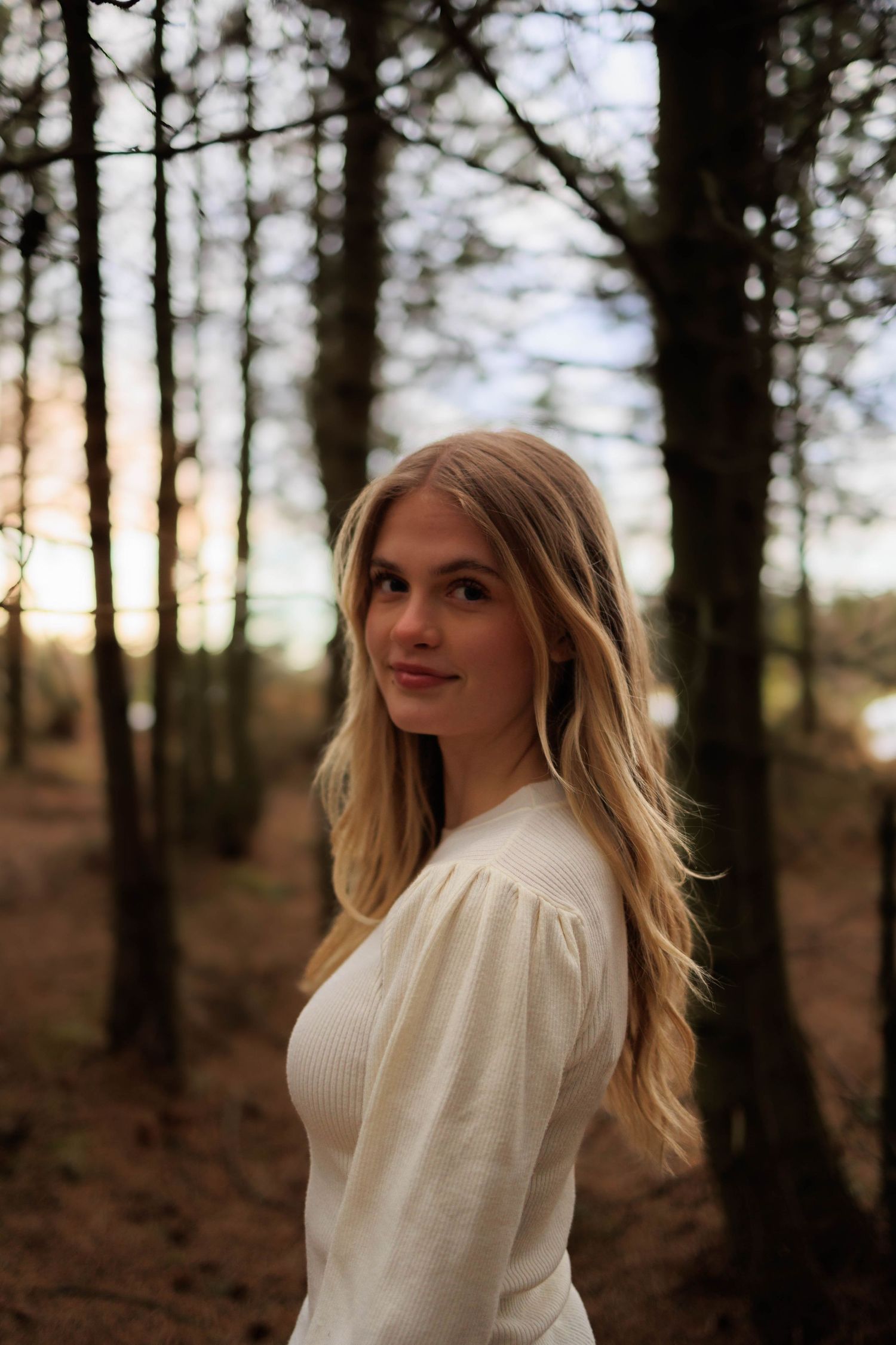 Portrait in white sweater against moody forest background at sunset.