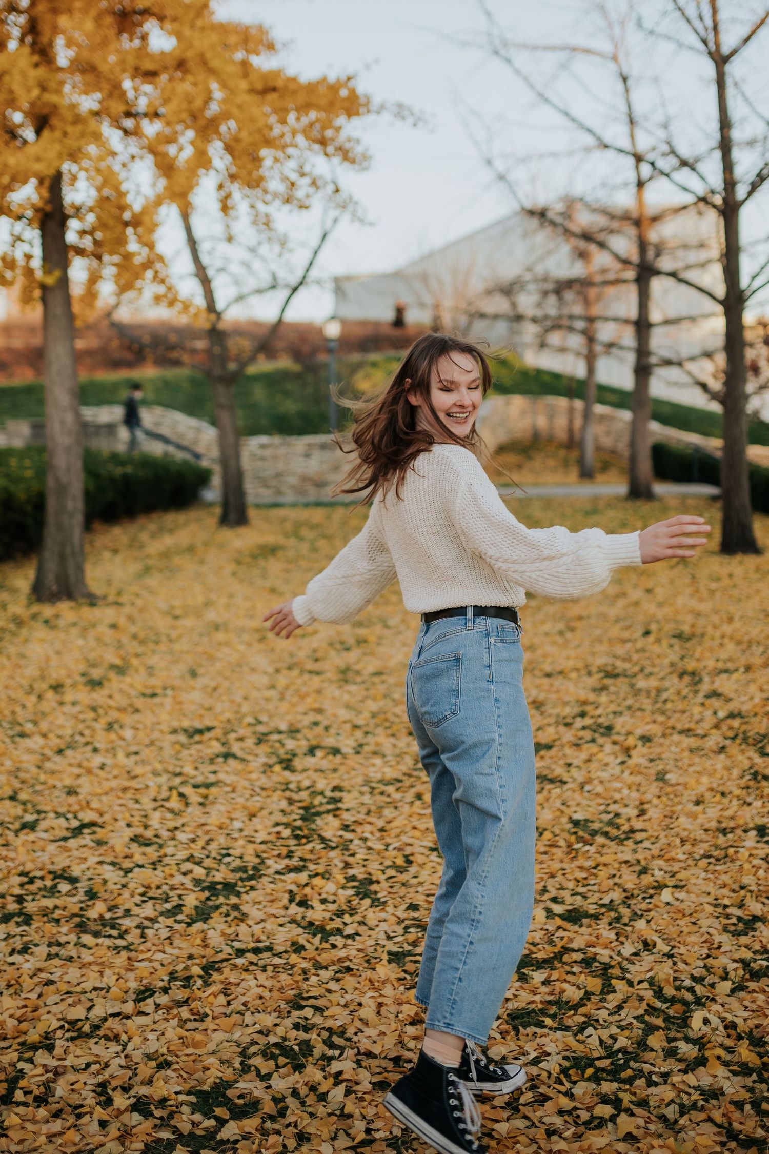 A high school senior twirls in an academic aesthetic outfit among bright yellow fall leaves during a senior photoshoot a