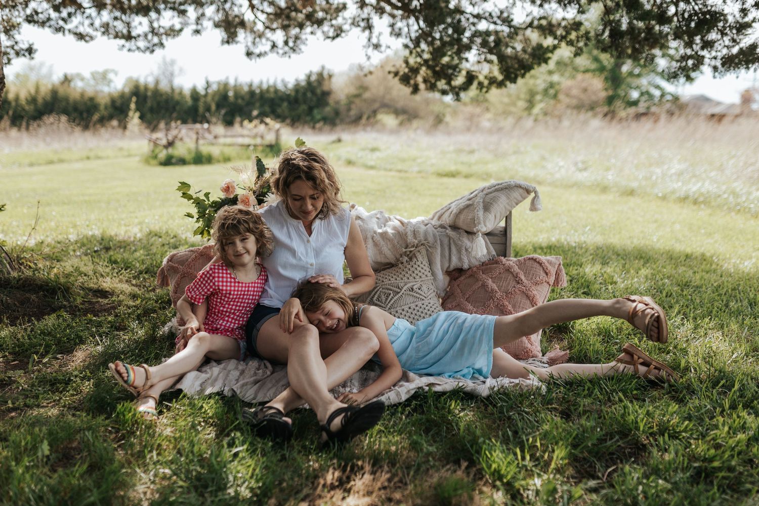 A mother and her two young daughters sit candidly on a boho blanket with pillows under a tree in an open field in Olathe