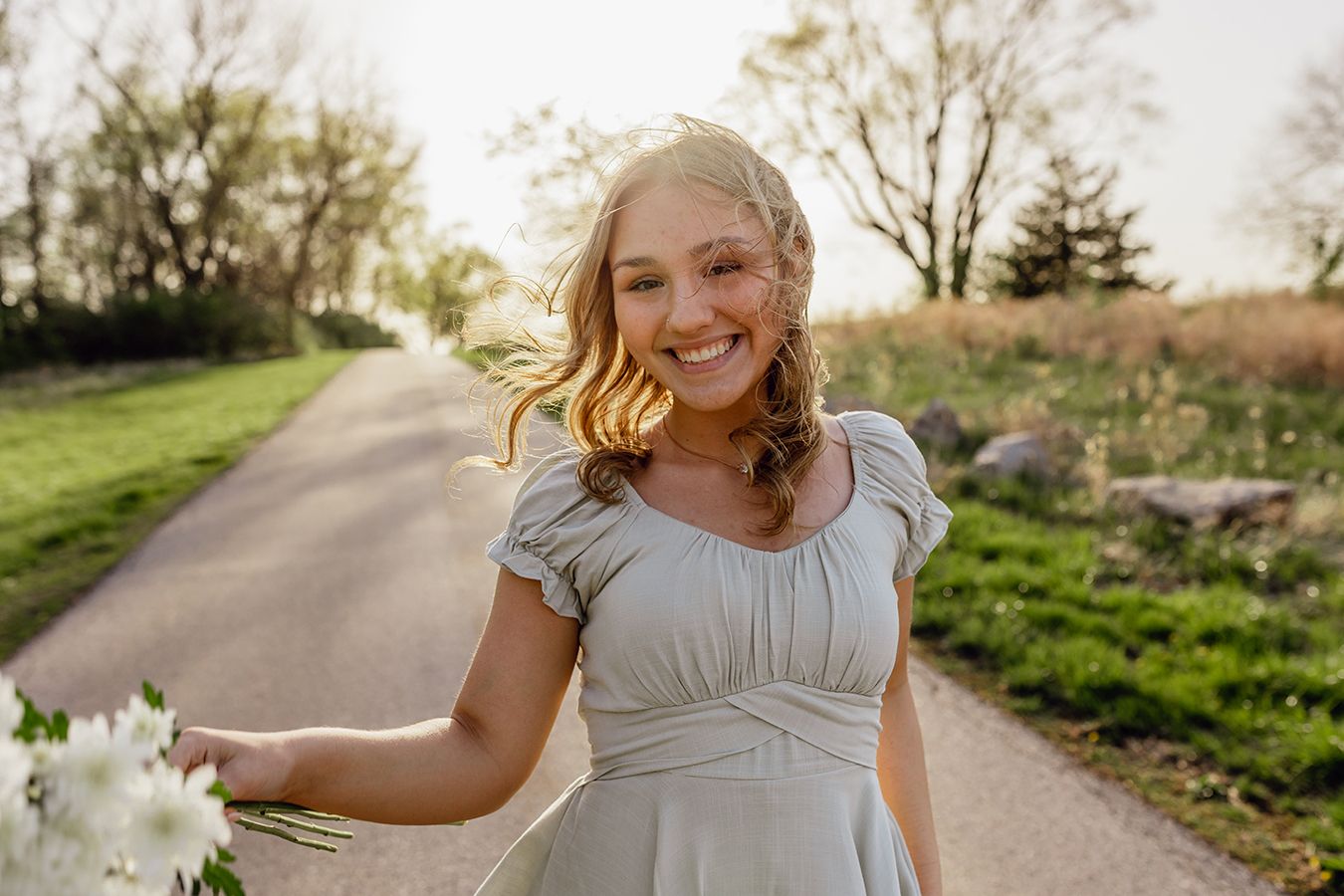 A high school senior smiles candidly while walking on a park trail with a white bouquet and wind in her hair in Martin C