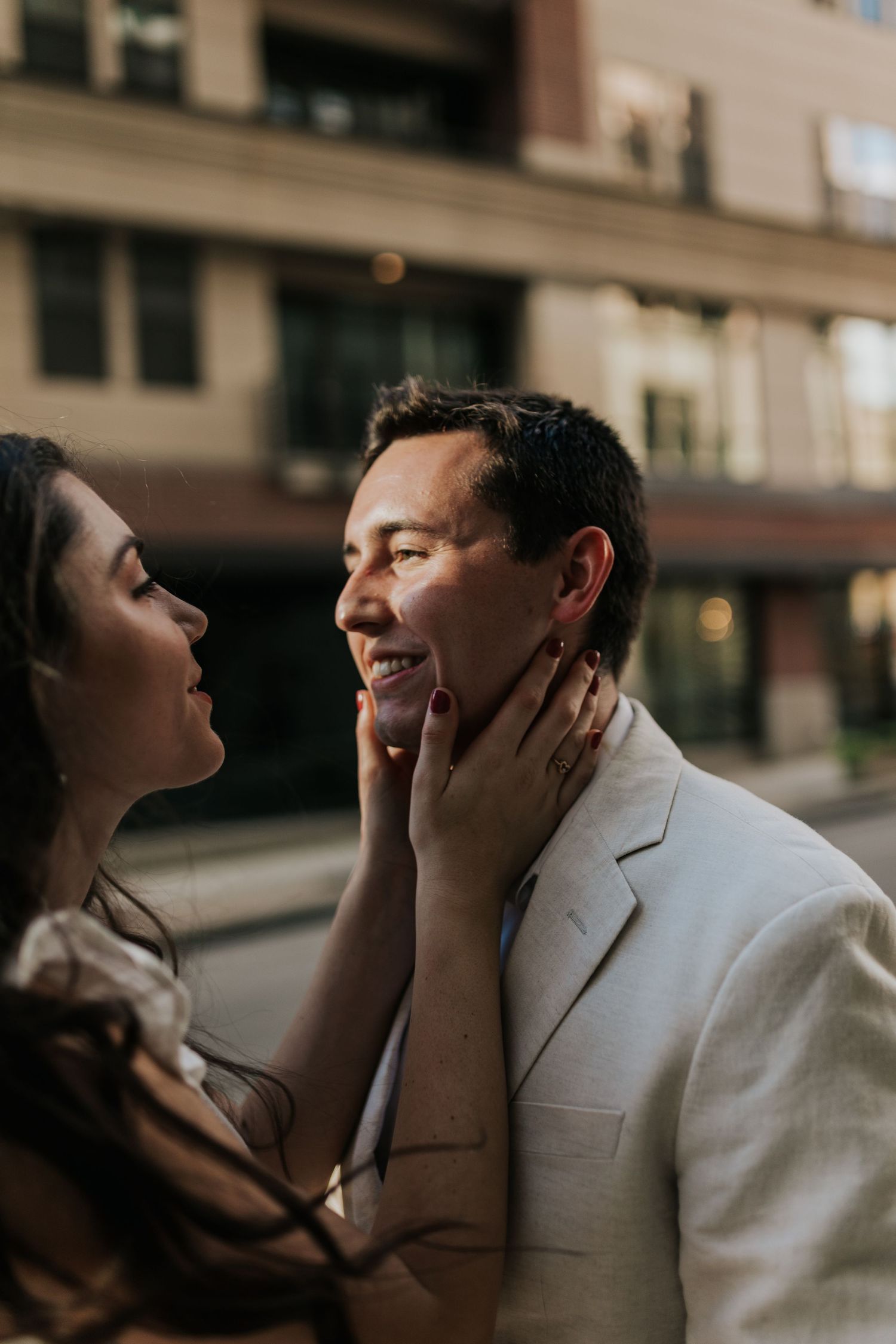 A brunette woman hold's her fiance's face as they smile candidly in neutral tones during an engagement photoshoot at gol