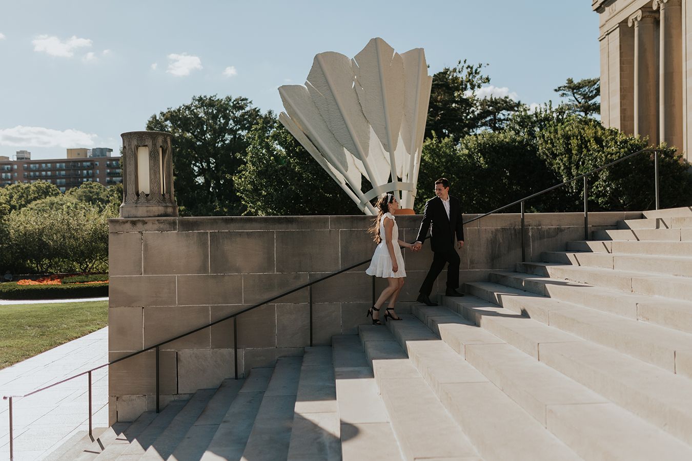 A newly engaged couple walk up the steps of the Nelson Atkins museum holding hands in front of the iconic shuttlecock in