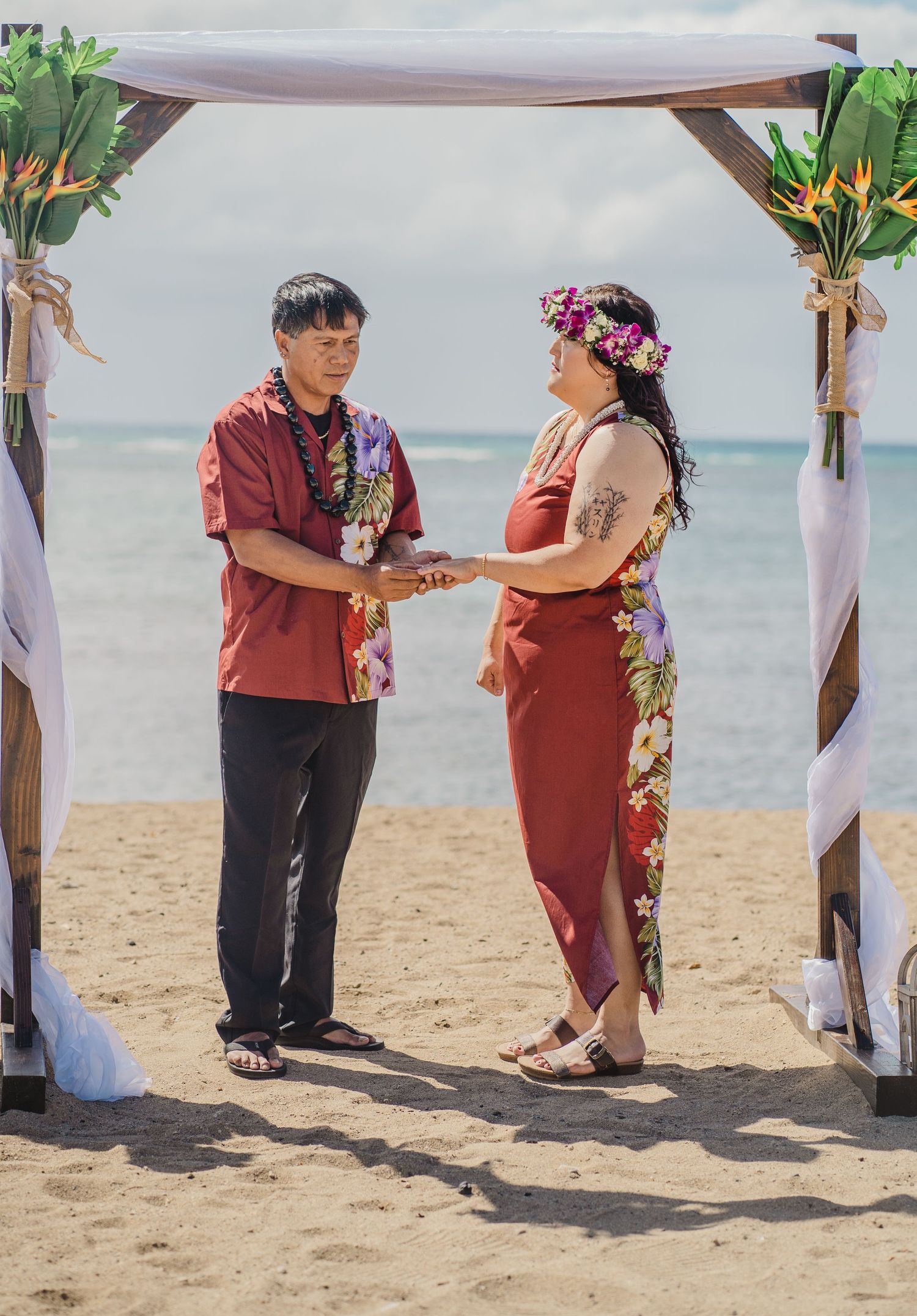 Traditional Hawaiian beach wedding ceremony under a wooden arch with tropical floral decorations.