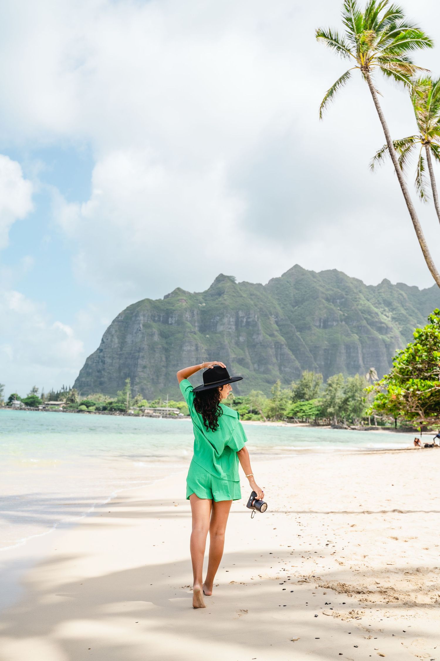 Woman in green walking along a Hawaiian beach with dramatic mountain landscape in background.