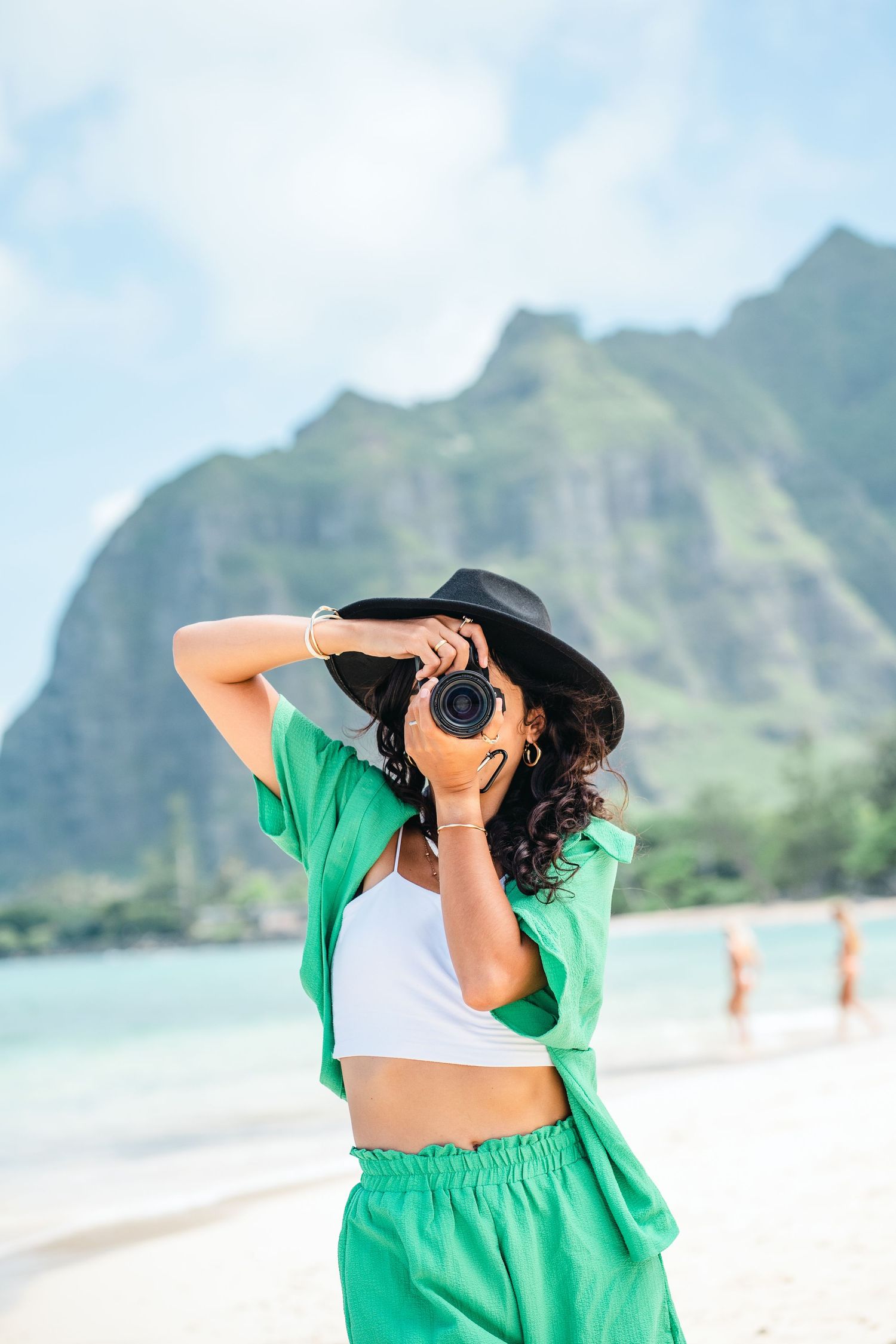 A photographer in green beachwear captures photos at a tropical beach with mountain backdrop.