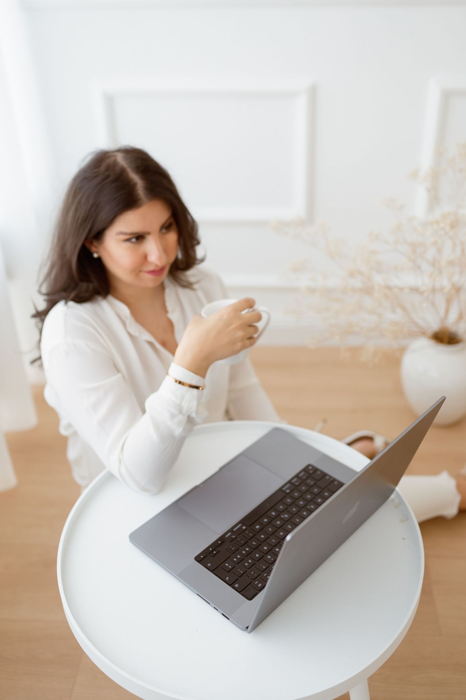 Woman in white outfit working on laptop at round table with coffee cup.