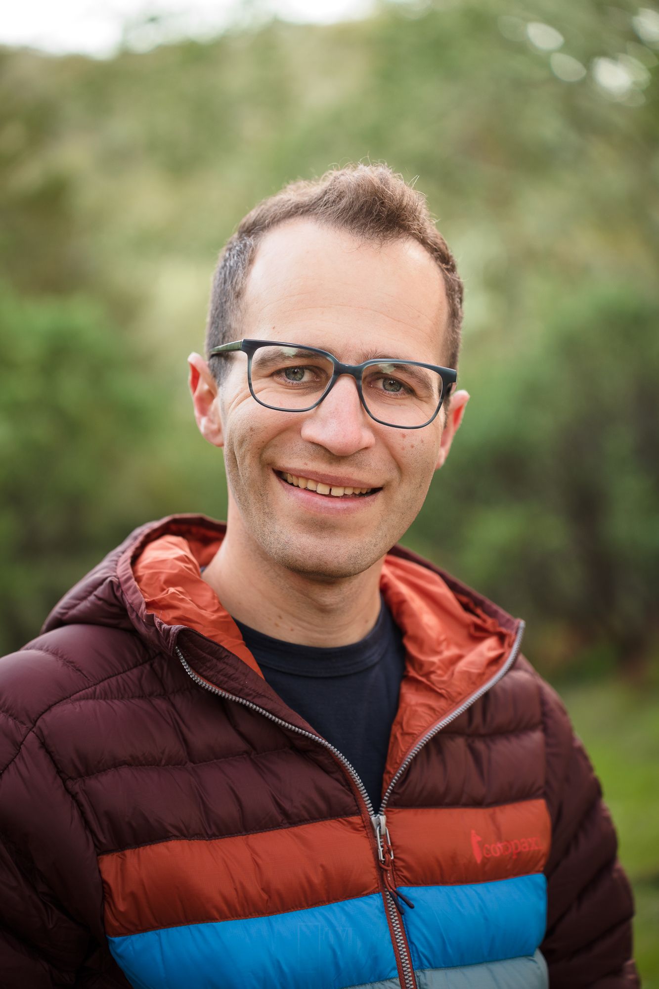 Person wearing glasses and a colorful striped puffer jacket smiles warmly against blurred outdoor background.