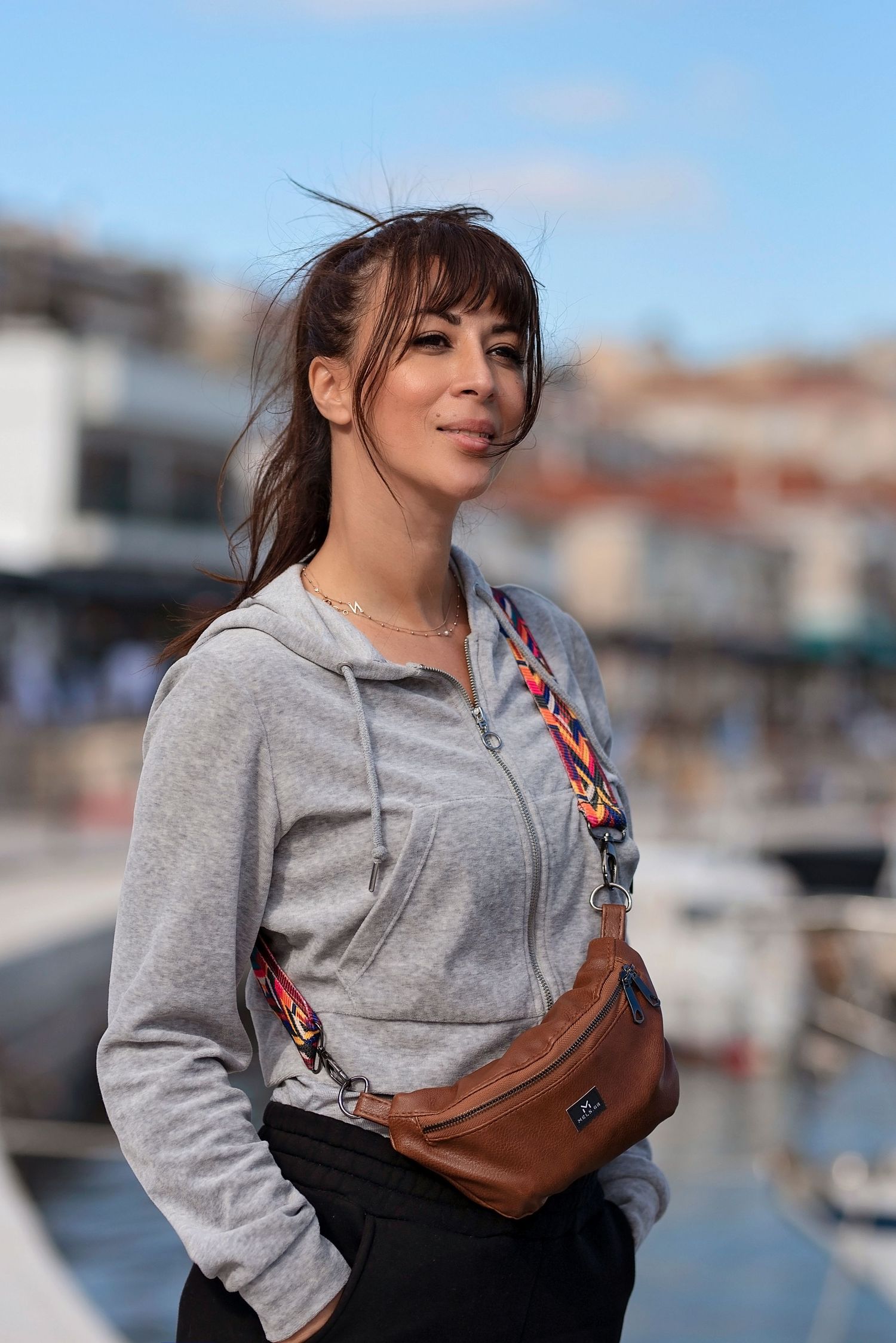 A woman in a gray hoodie and brown crossbody bag stands near a marina on a sunny day.