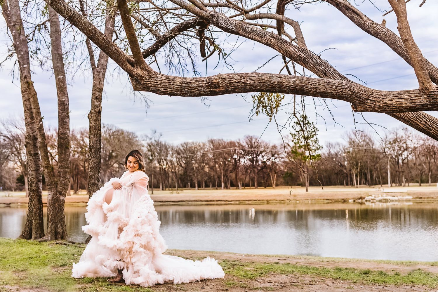 Blush wedding gown photographed by serene lake with overhanging tree branches.