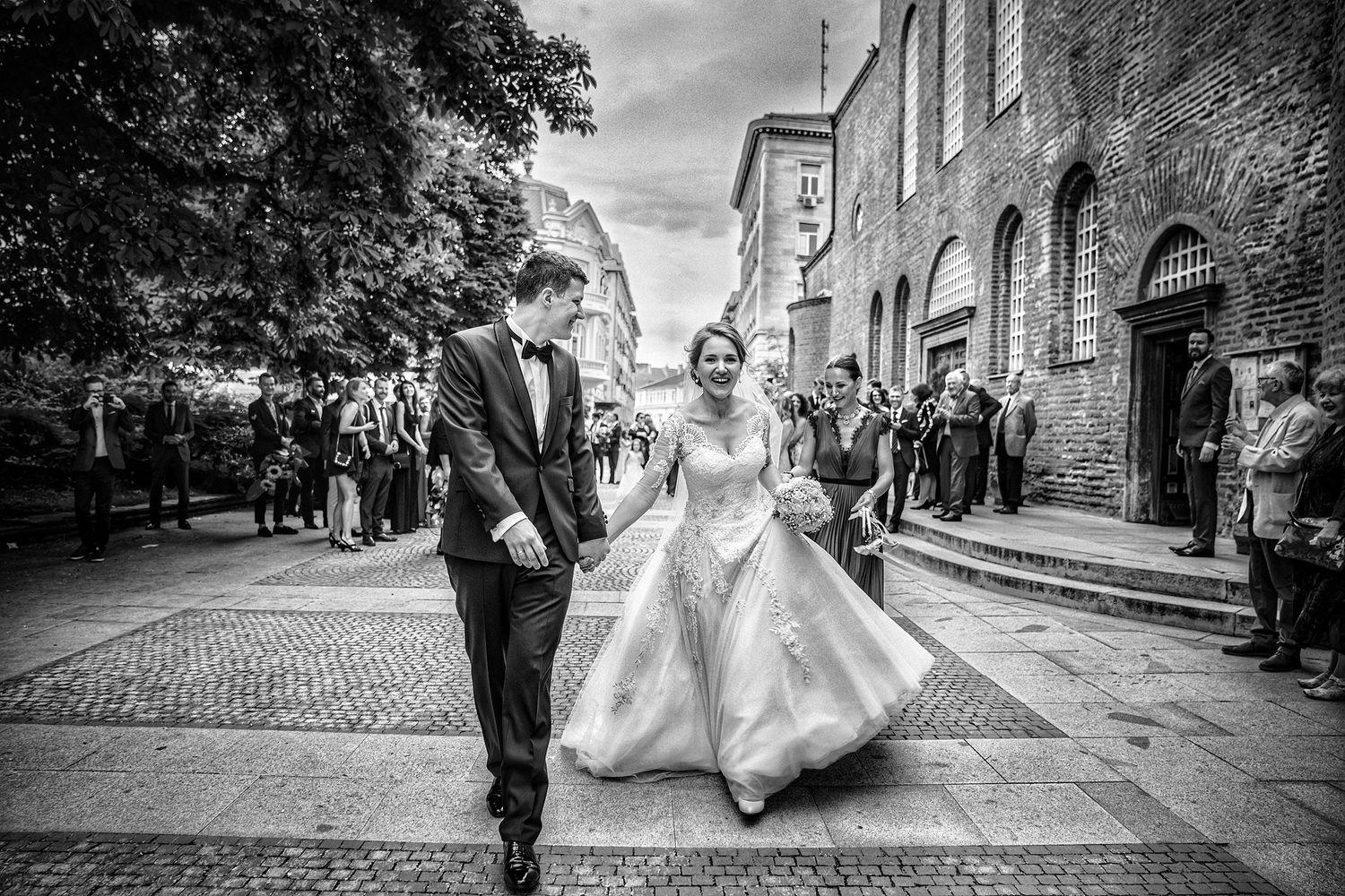 A couple walks joyfully down a cobblestone street while onlookers gather on a bright wedding day.
