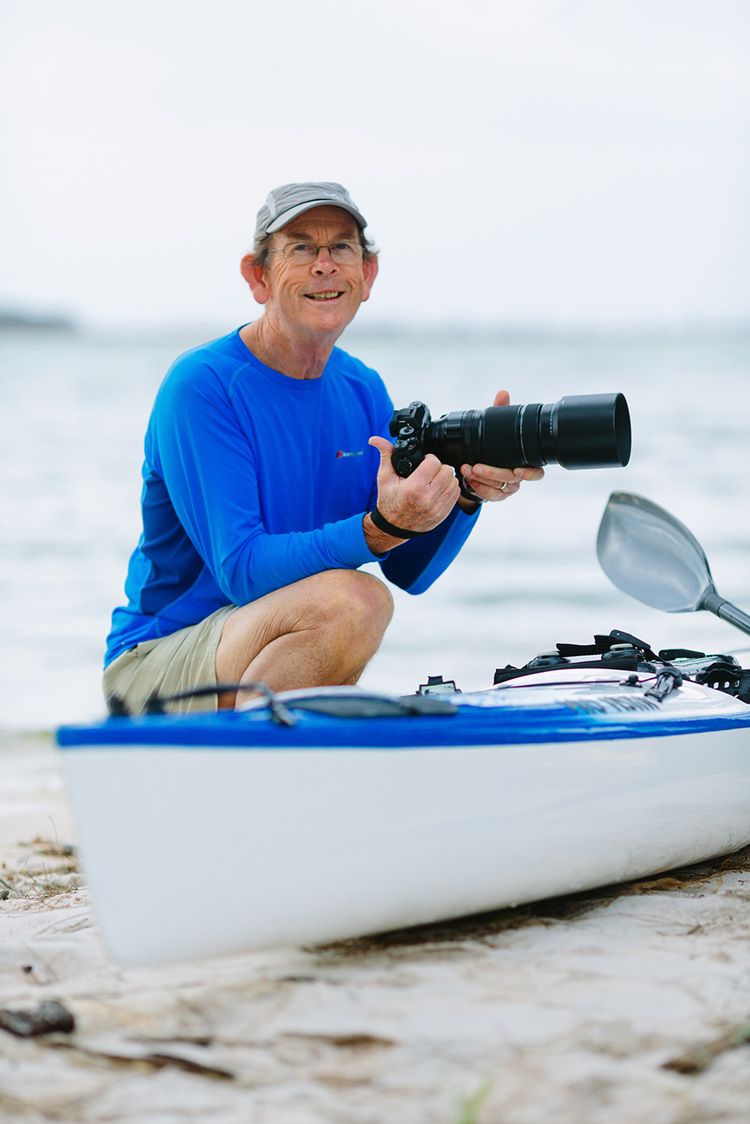 Photographer in blue shirt with camera gear sitting beside kayak on beach.