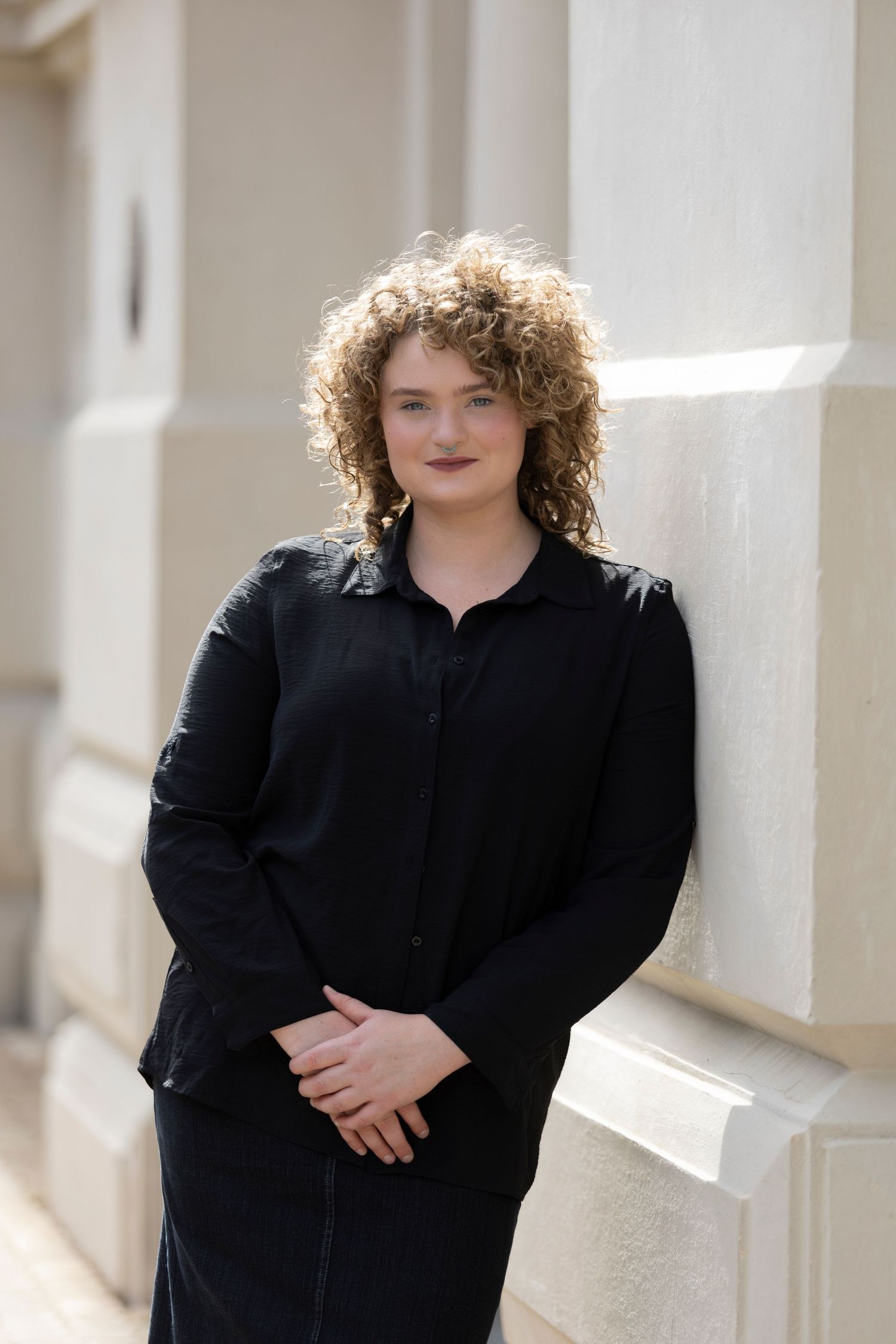 Portrait of someone with curly hair wearing all black against a white building exterior.
