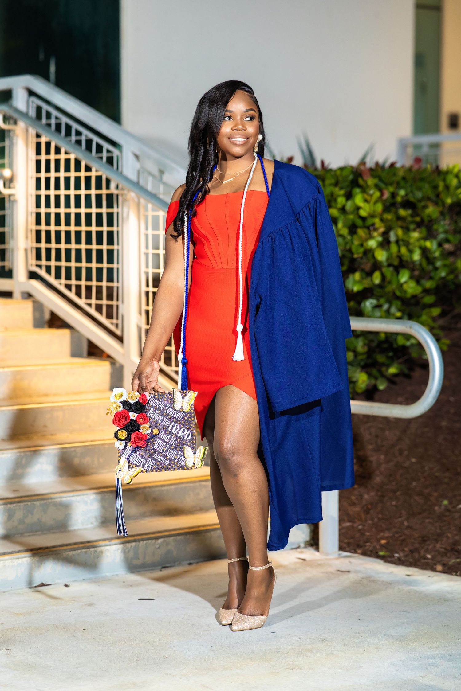 Graduate in red dress and blue graduation gown holds decorated cap on outdoor steps at night.