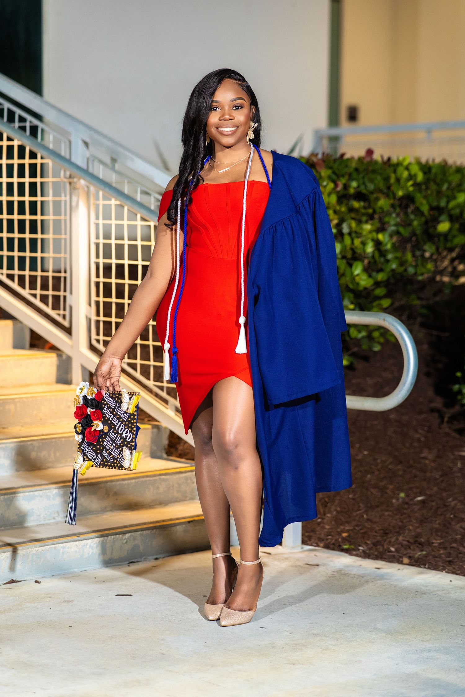 Smiling graduate in red dress with blue graduation gown walks down steps holding decorated cap.