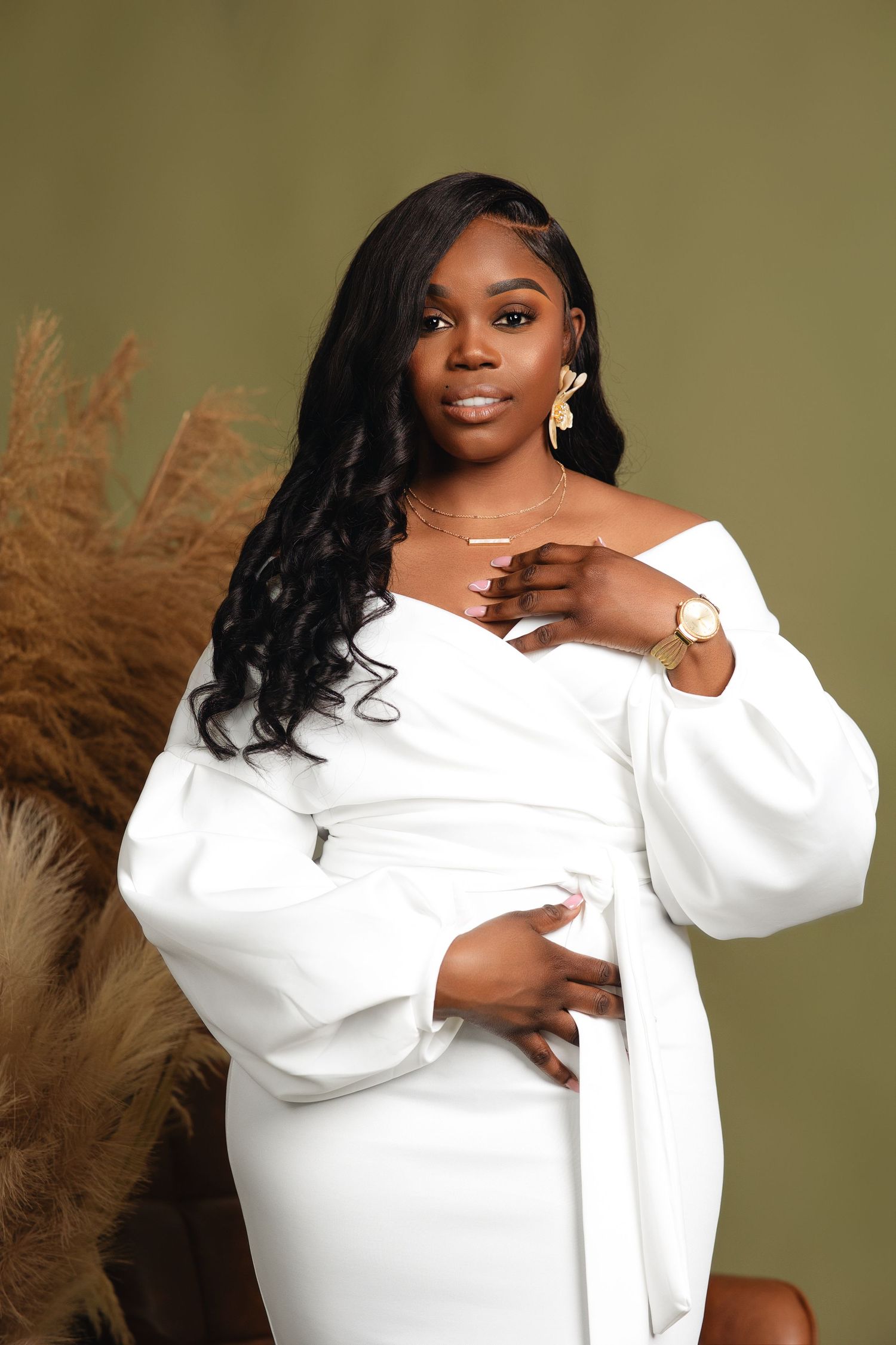 Woman in white off-shoulder wrap dress poses with gold jewelry beside pampas grass on olive background.