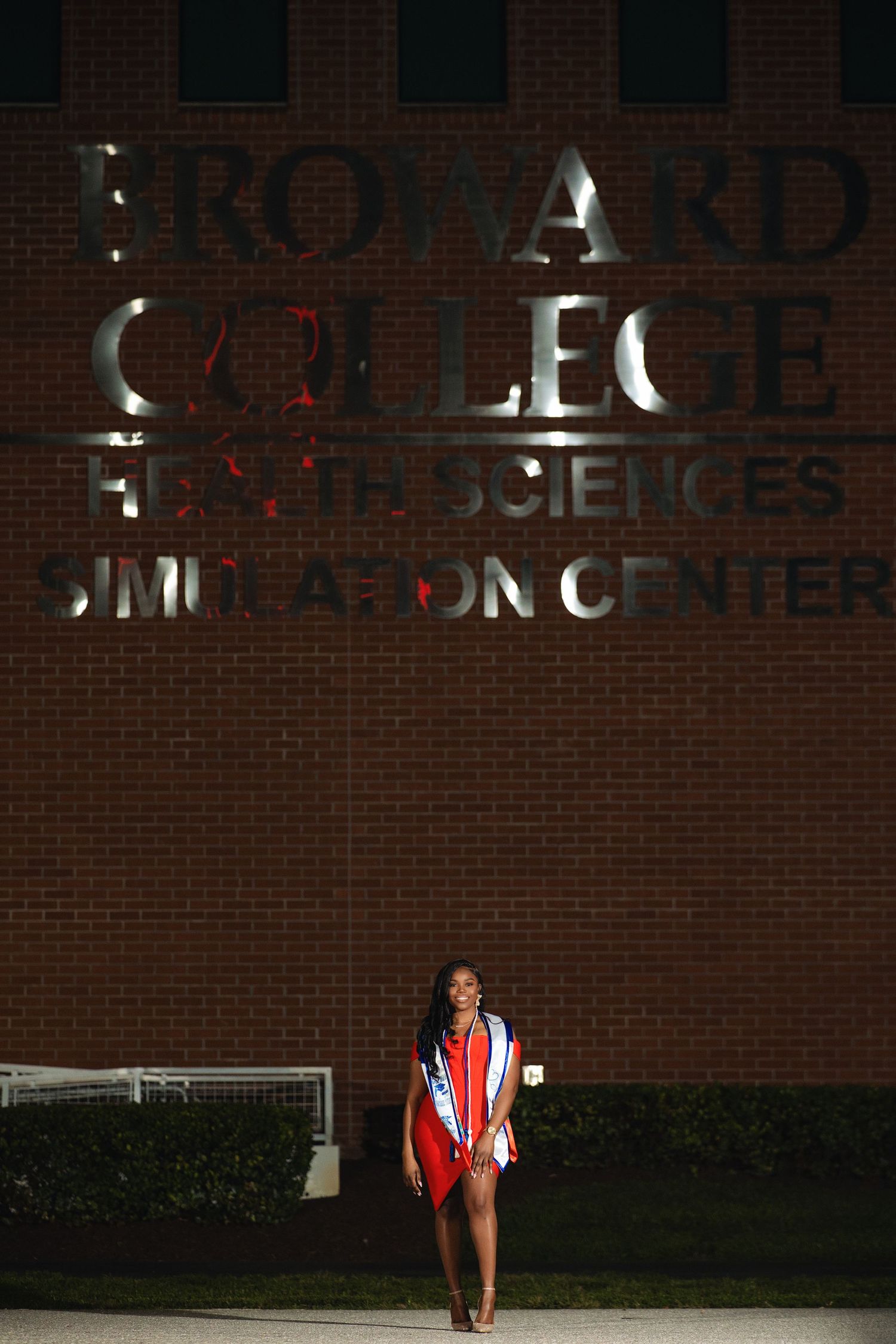 Graduate poses at night in front of Broward College Health Sciences Simulation Center brick building.