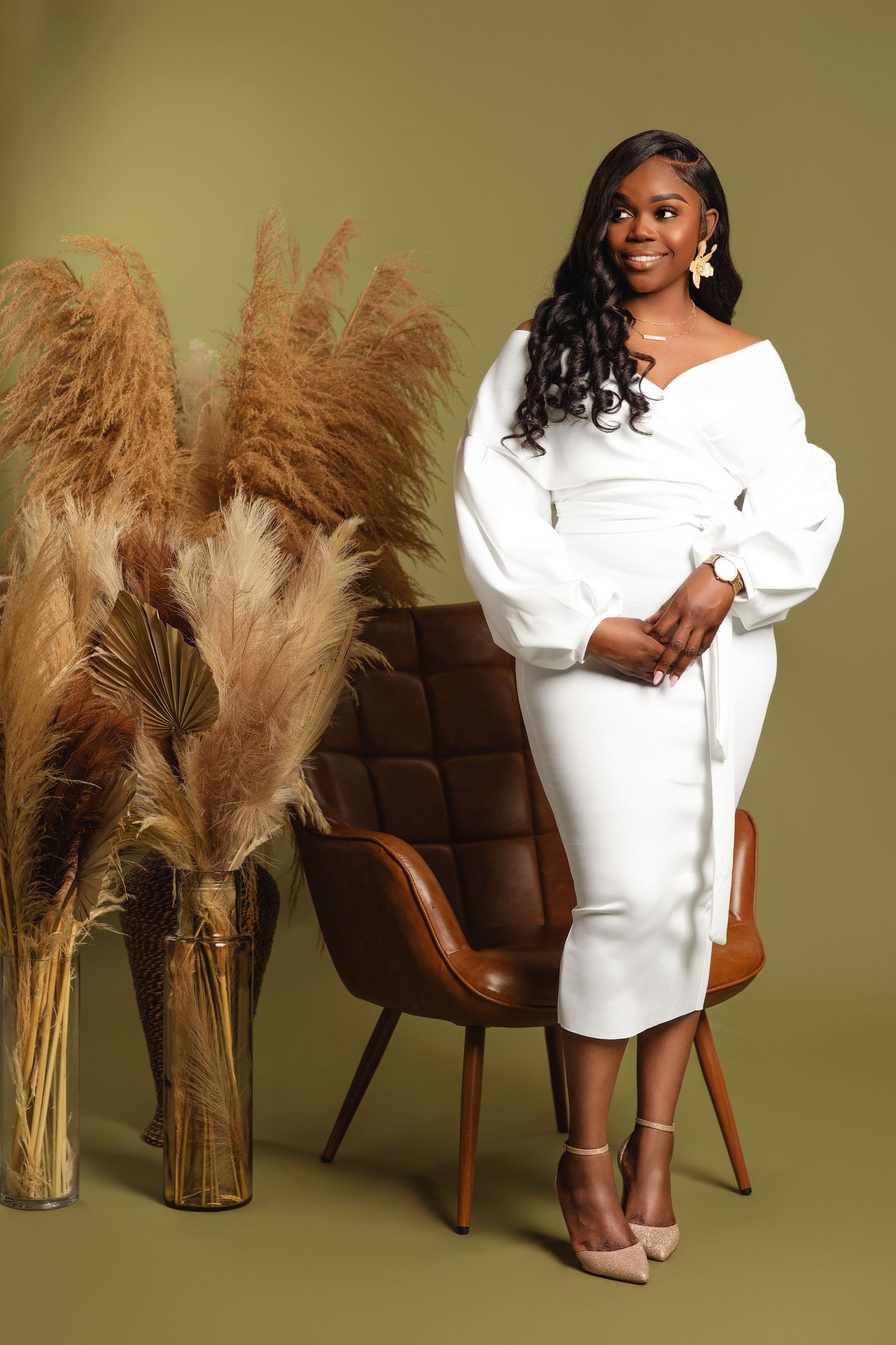 Woman in white off-shoulder midi dress poses elegantly beside pampas grass decor on olive green background.
