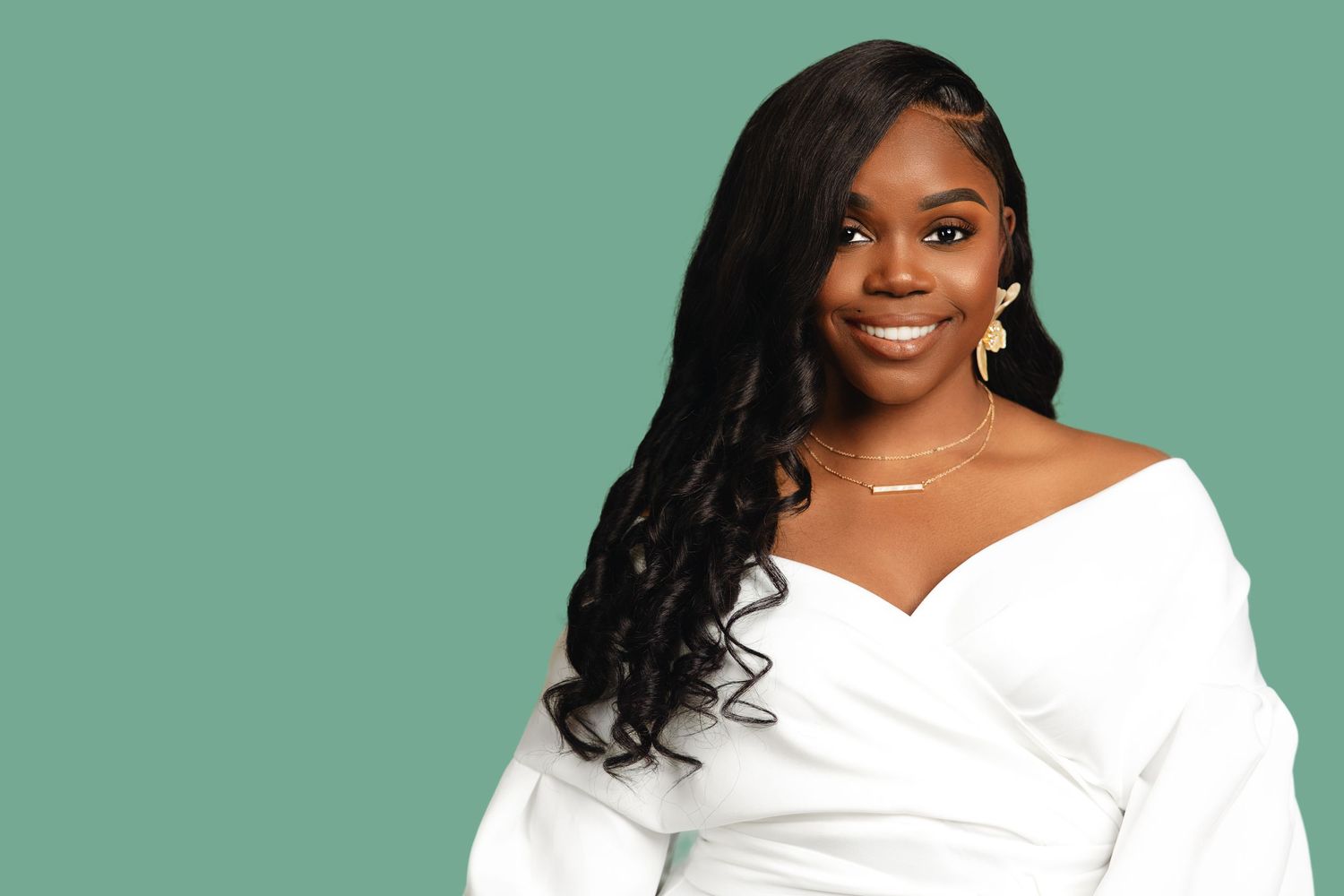 Smiling woman in white off-shoulder top poses against teal green background with long black hair.