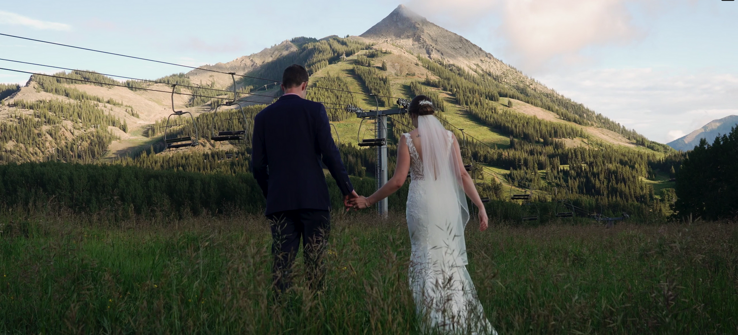 A couple walking hand in hand through a grassy meadow with a majestic mountain peak in the background at sunset.