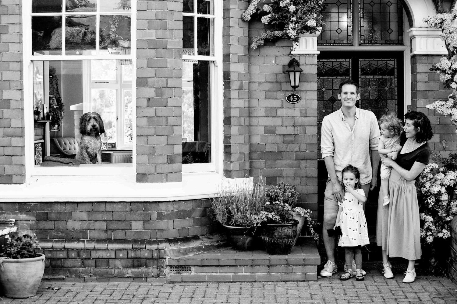 A young family stand in front of their home watched by their family dog who stand in the large bay window
