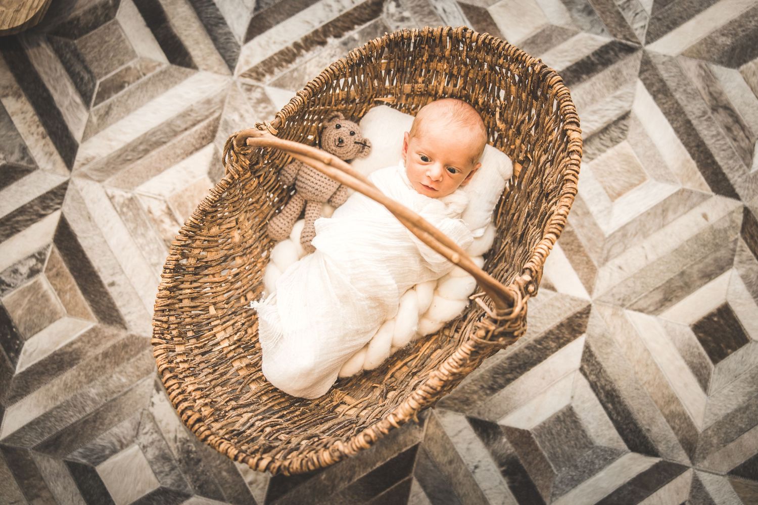 A sleeping infant wrapped in white swaddle lies in a woven wicker moses basket on a geometric patterned floor.  Houston Newborn Photographer