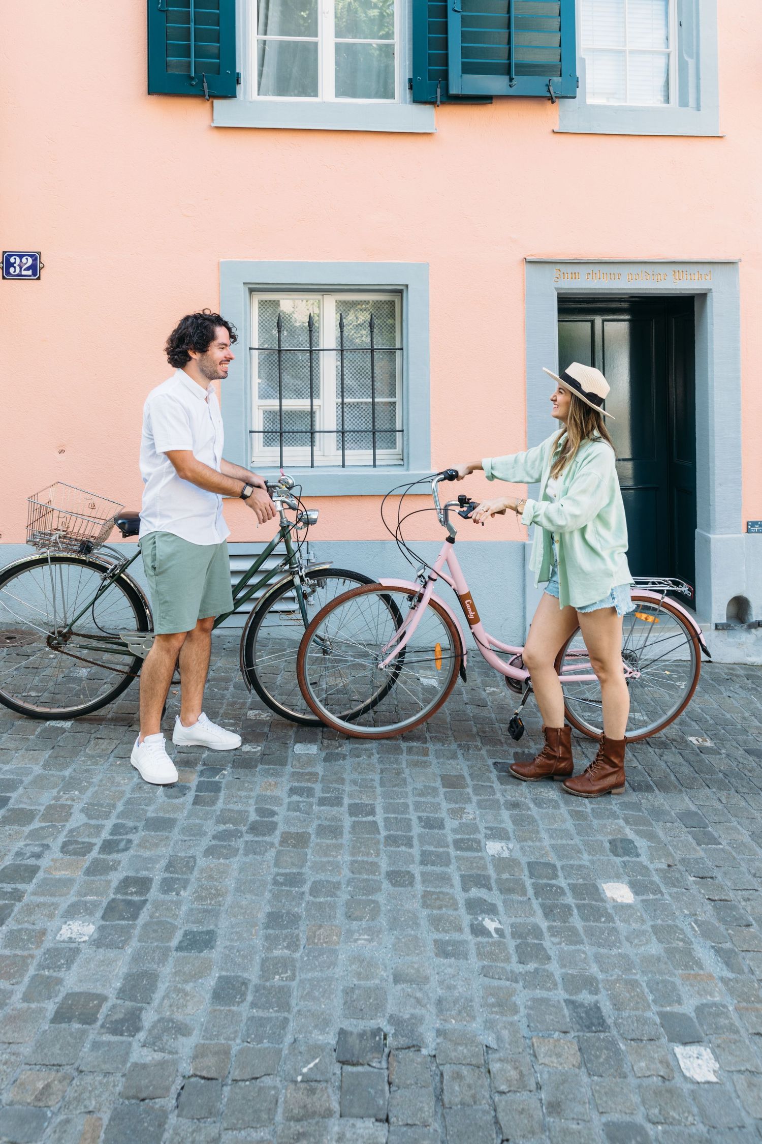 Two people with bicycles chatting on cobblestone street in front of pastel pink building.