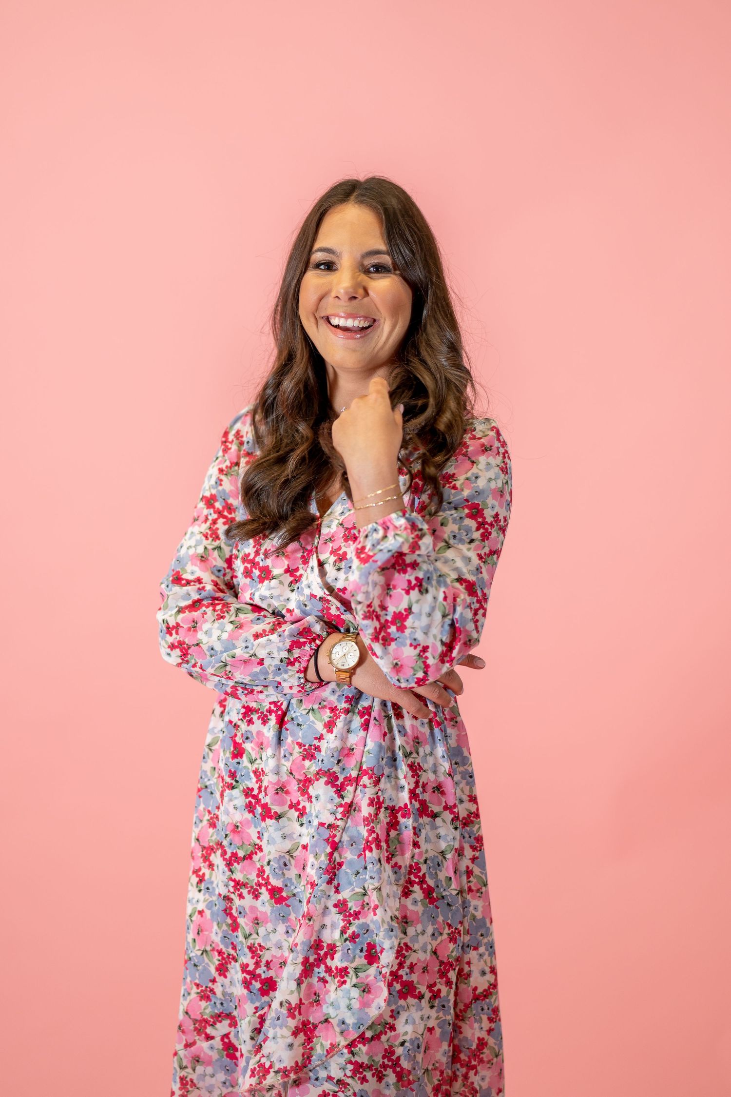 Portrait against pink background wearing a floral print dress with long wavy hair.