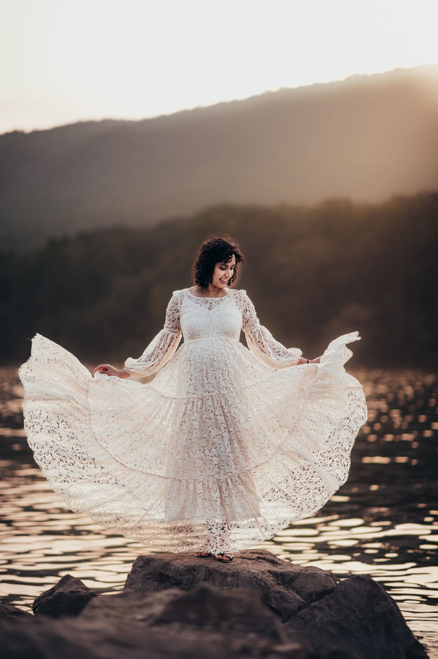 Woman in white lace dress twirling on rocks by water at sunset.