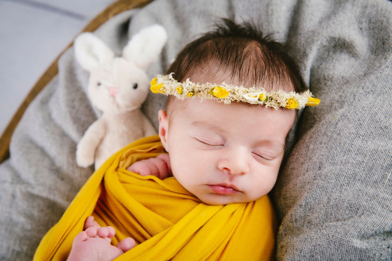 Sleeping baby wearing yellow wrap and floral crown with white stuffed bunny toy nearby.