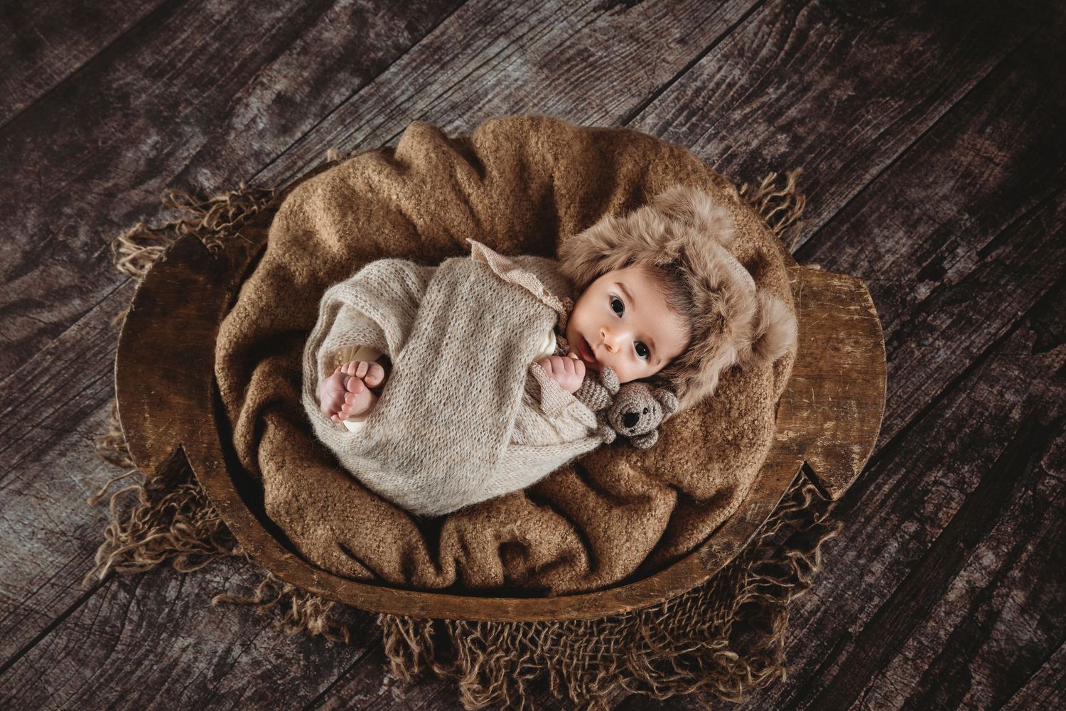 Newborn wrapped in gray swaddle lying in wooden bowl on dark wooden floor.