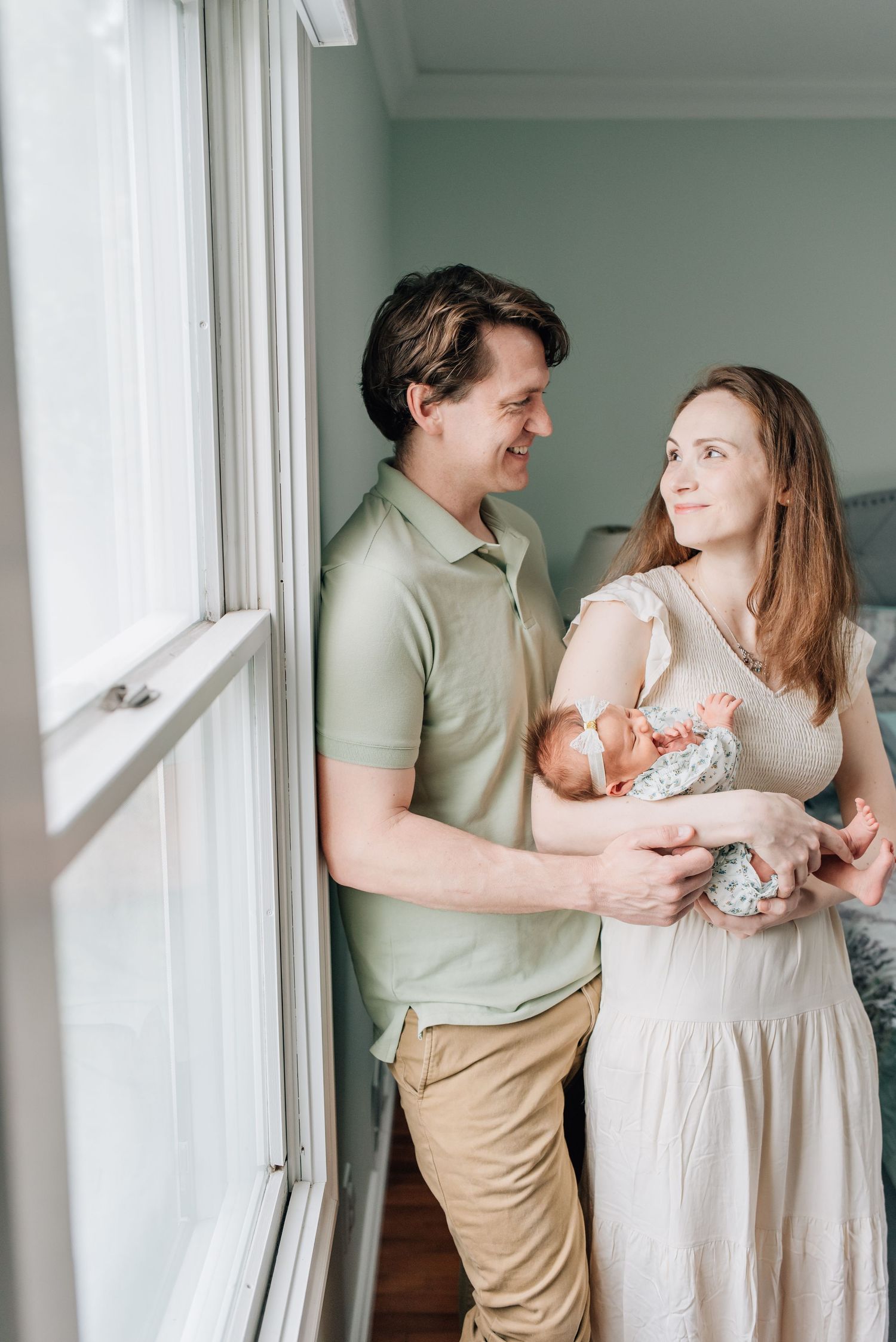 A man in a green polo sheet holds his wife close as they smile with their newborn baby girl.
