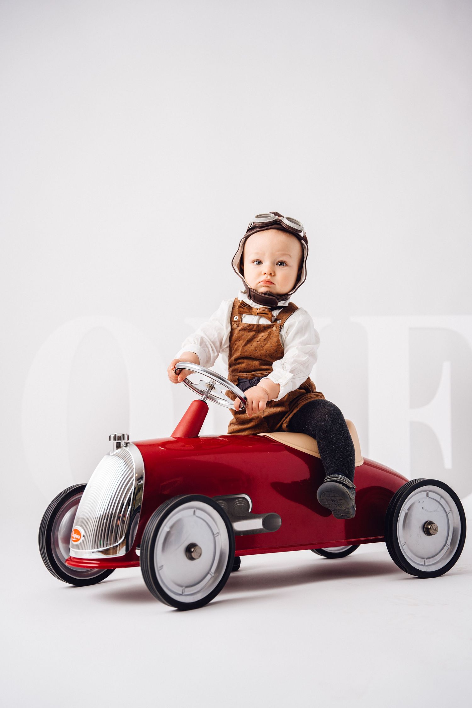 Baby in vintage clothing riding a red pedal car toy against white background.