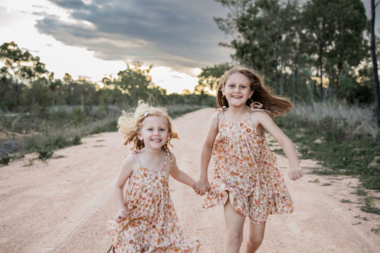 Two girls in matching floral dresses run joyfully down a dirt path.