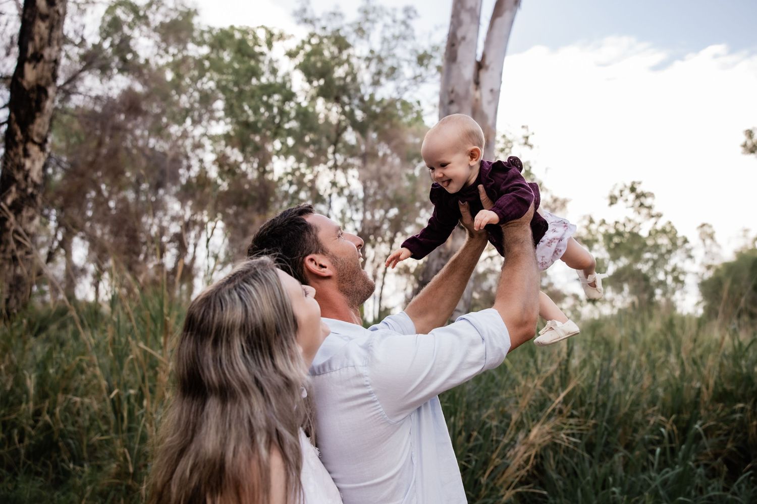 Parents lifting baby in the air against backdrop of tall grass and eucalyptus trees.