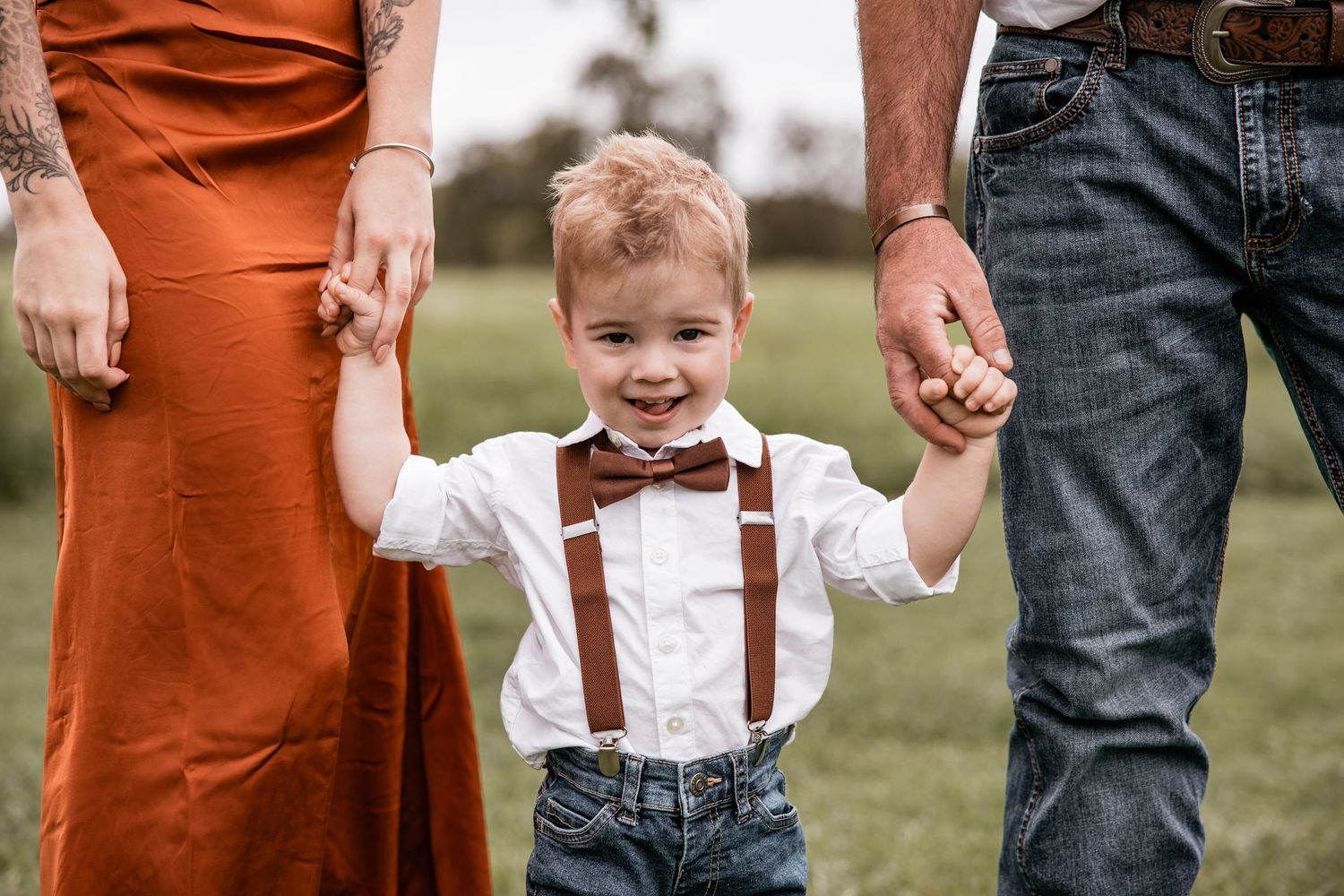 Small child in suspenders and bow tie holds hands with parents on grassy field.