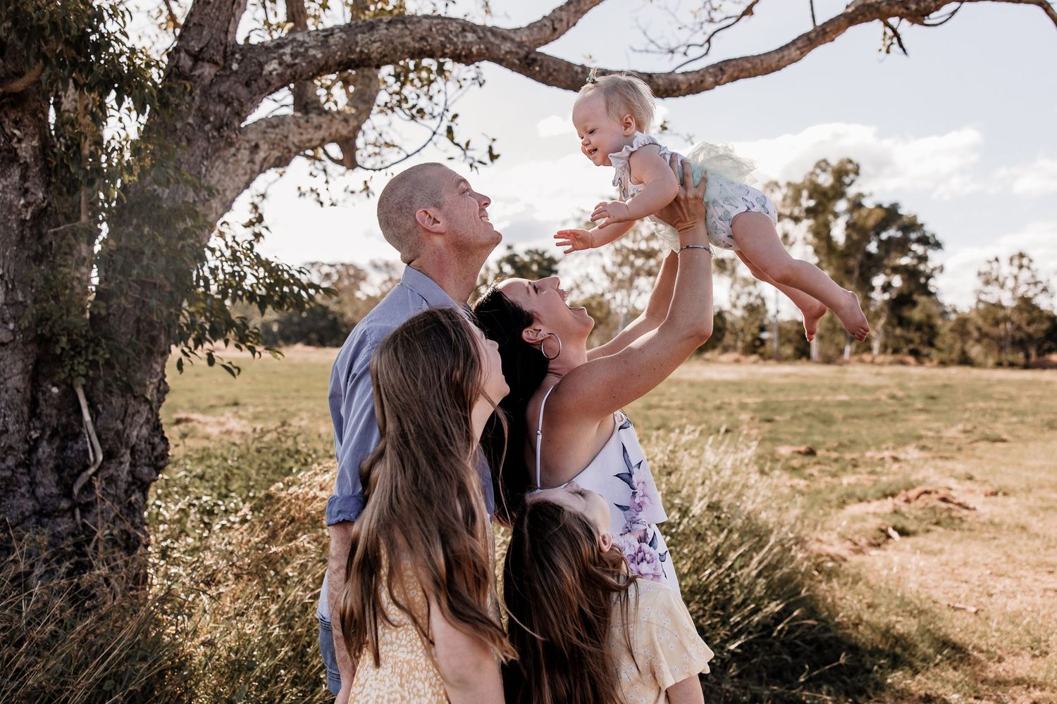 A joyful family moment as parents lift their baby in the air under a large tree.