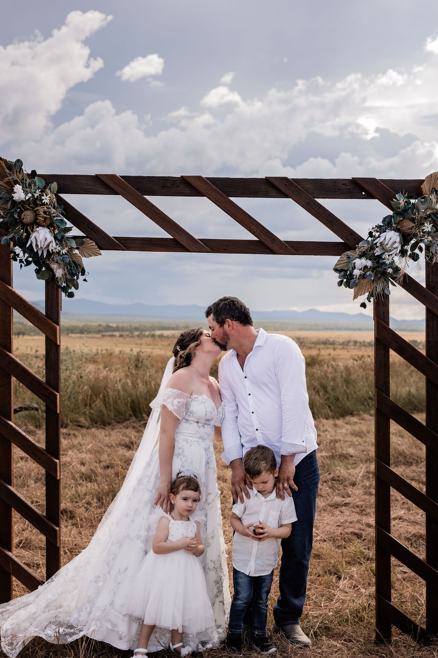Family gathered under wooden wedding arch in rustic outdoor setting.