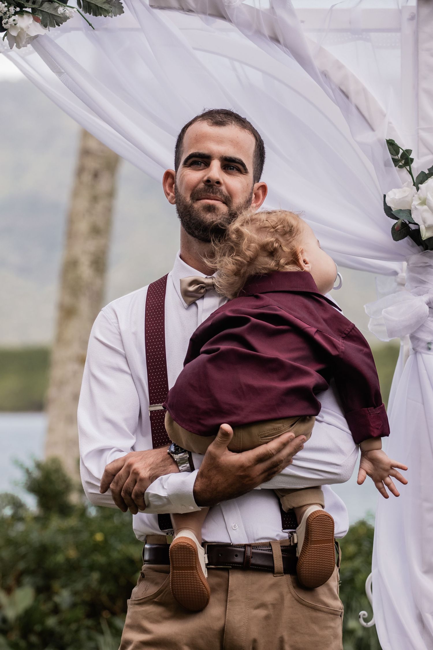 Person in formal attire holding small child at outdoor wedding.