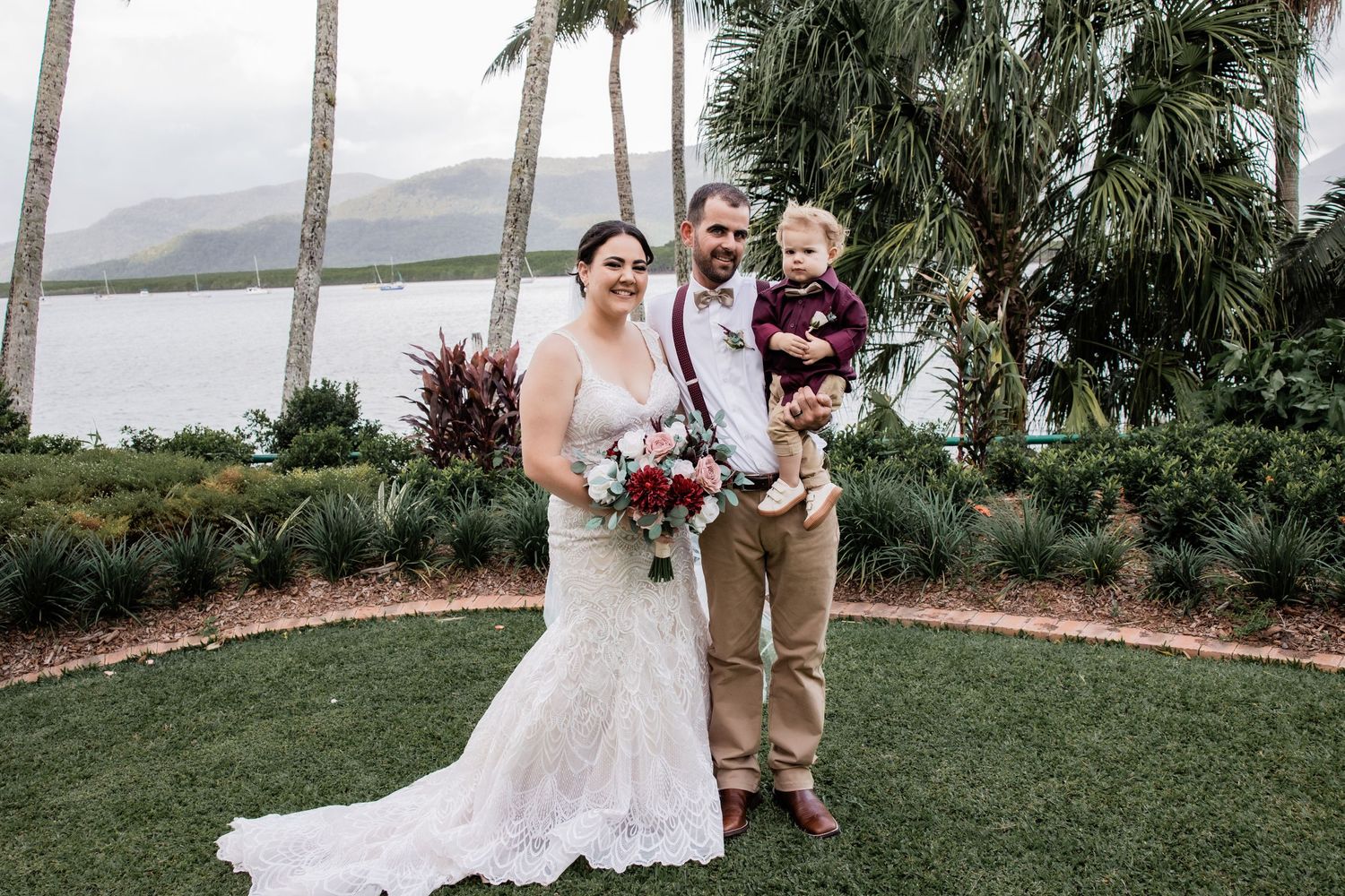 Newlyweds pose with child by tropical waterfront in formal wedding attire.