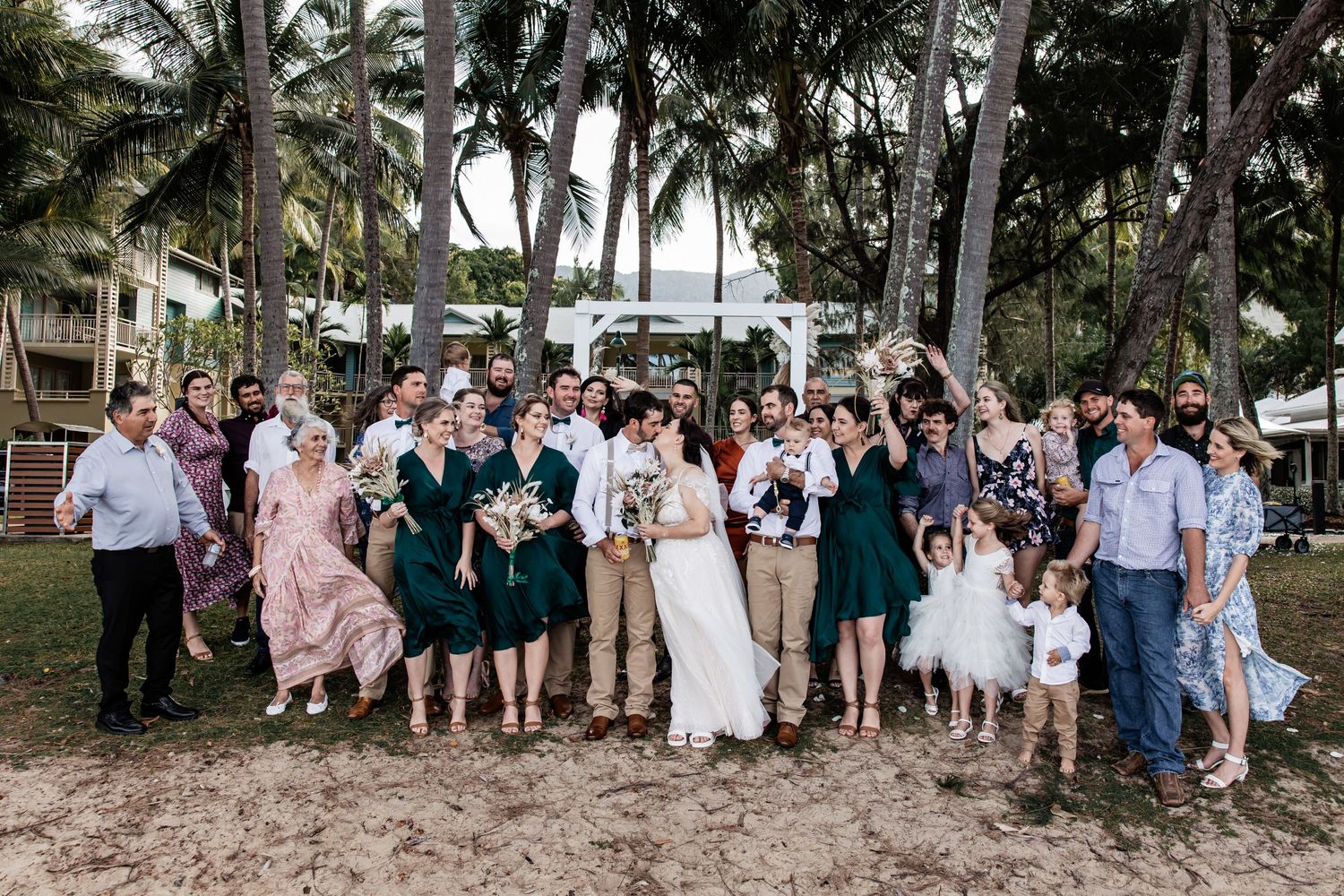 Wedding group photo with palm trees and ocean backdrop in tropical setting.