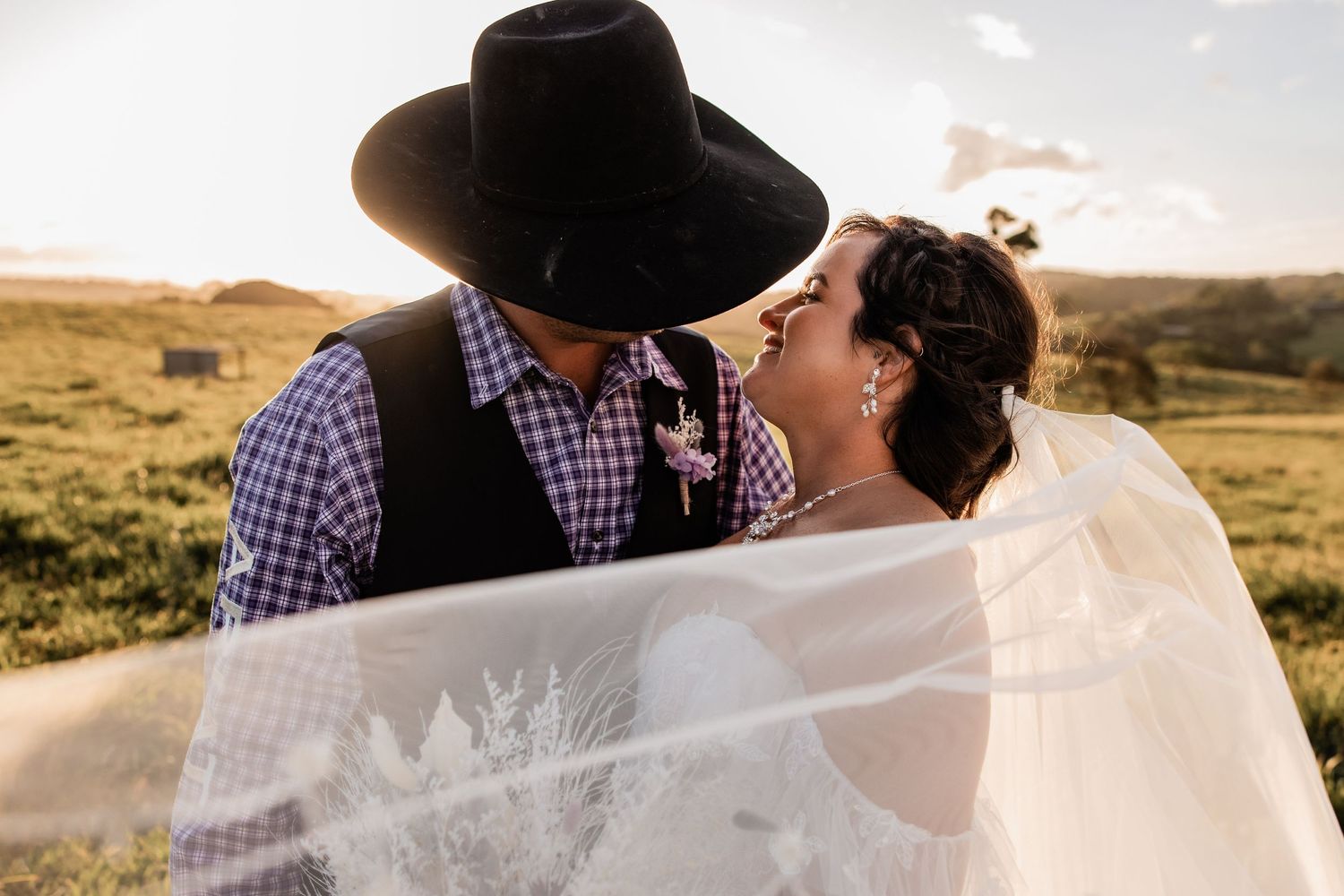 A romantic wedding couple embraces in a field at sunset with the bride's veil flowing in the breeze.