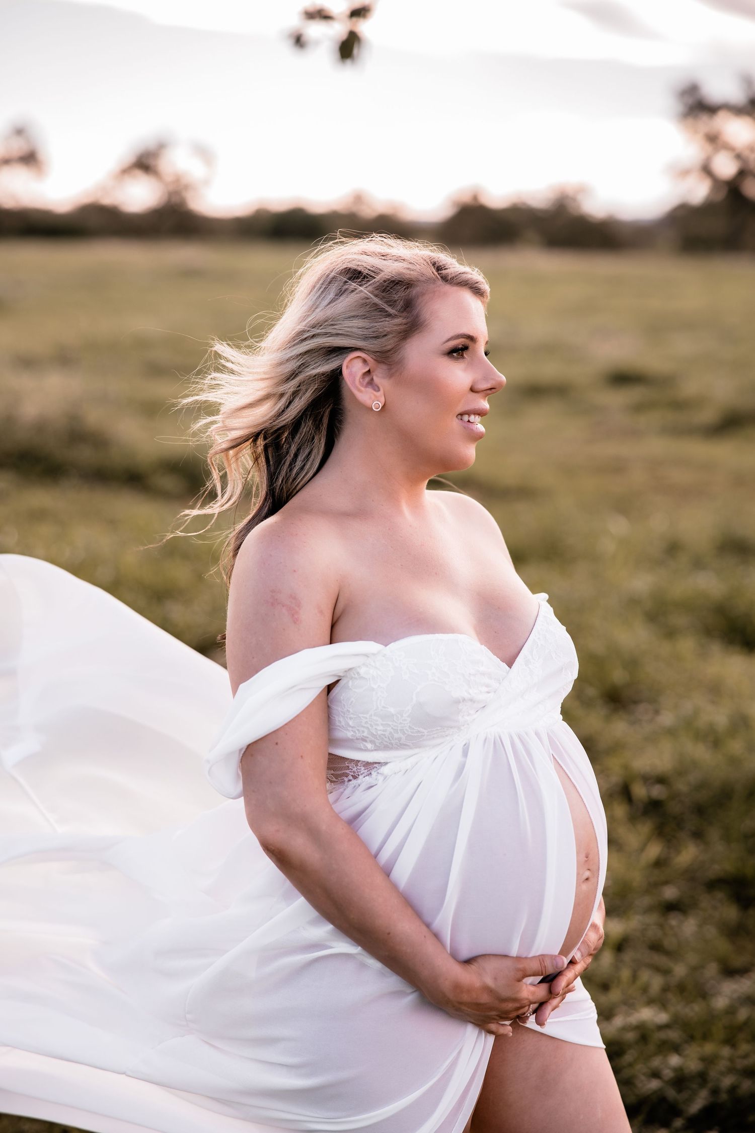 Expectant mother in flowing white dress poses in field at sunset.