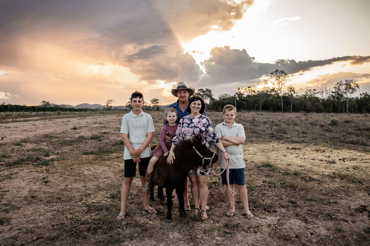 Family stands with donkey in field during dramatic sunset with storm clouds.