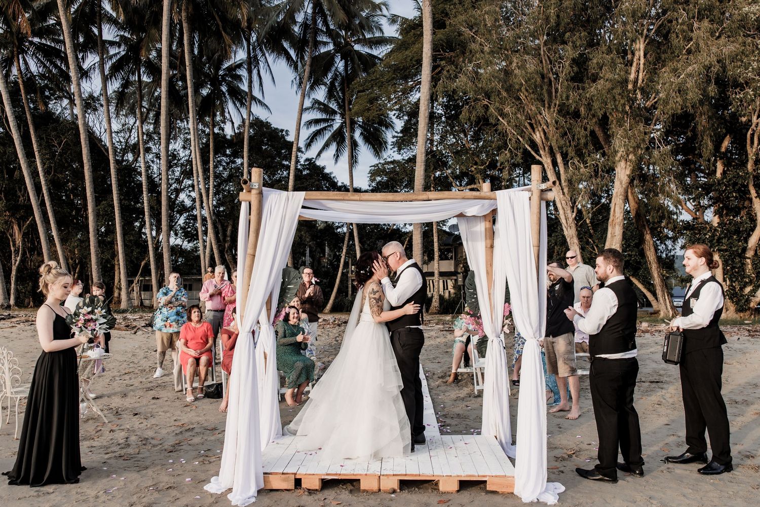 Beach wedding ceremony under white draped arch with palm trees in background.