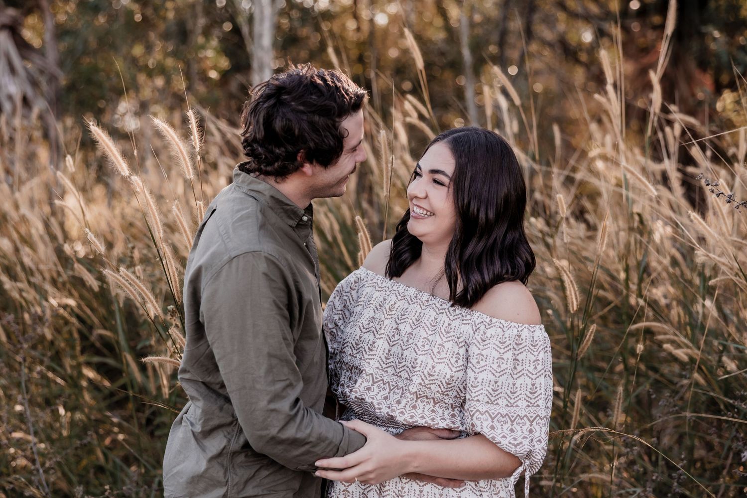 A romantic couple shares an intimate moment while standing in tall grass during a sunset photoshoot.