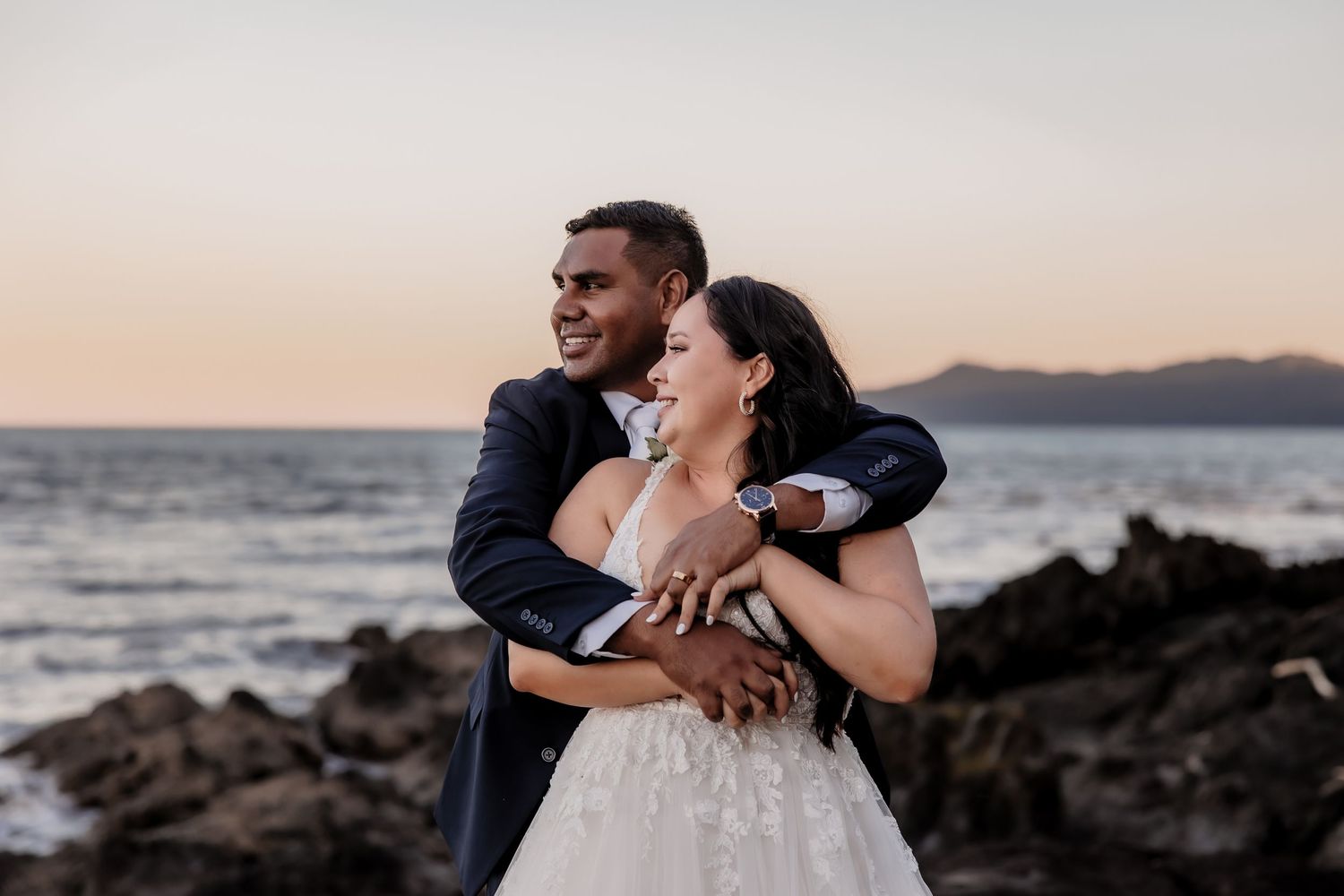 Wedding couple embraces on rocky coastline at sunset.