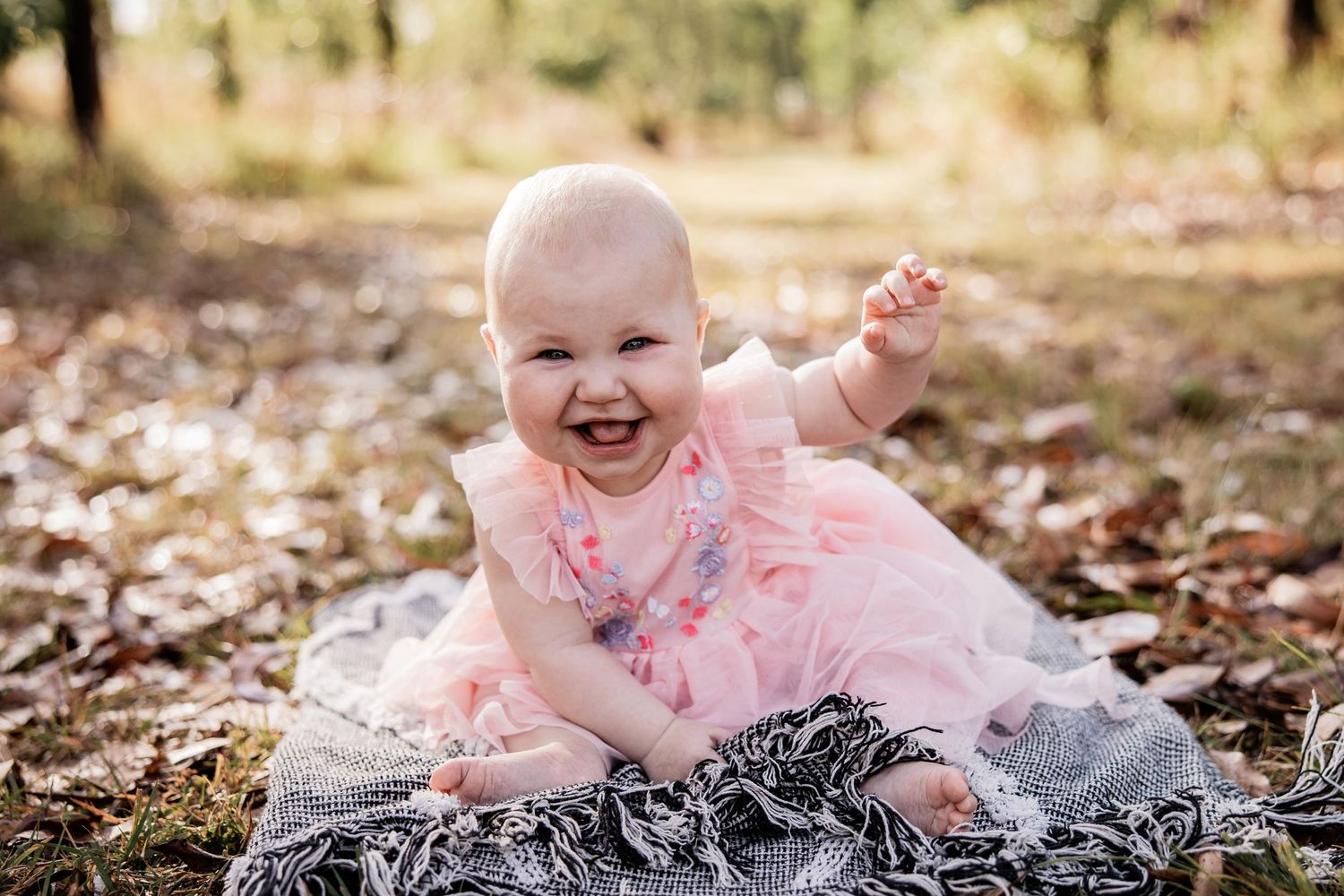 A joyful baby in a pink tutu dress laughs while sitting on a blanket outdoors.