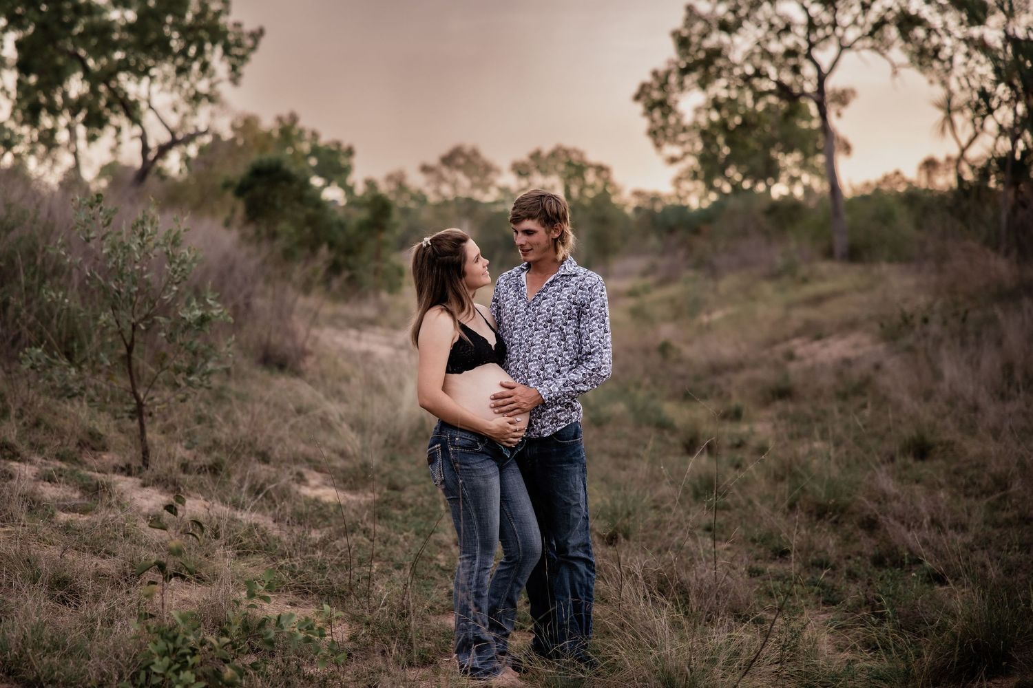 A romantic couple embraces on a hillside at sunset surrounded by natural terrain.