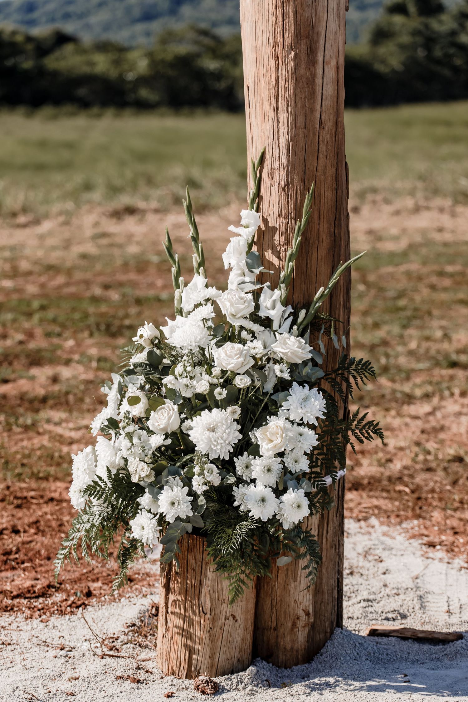 White floral arrangement with greenery displayed on wooden post outdoors.